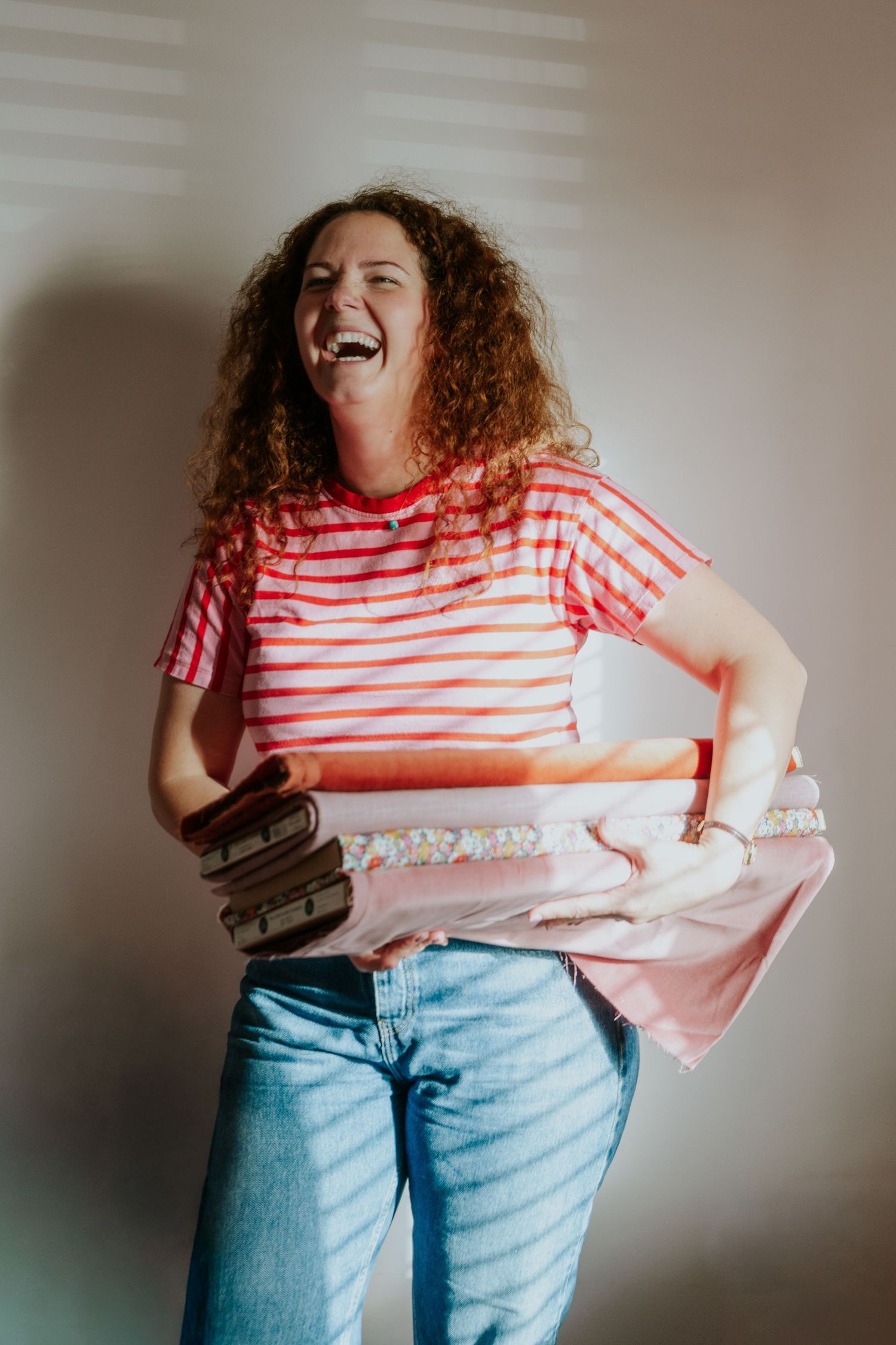 Person in red and white striped shirt and jeans laughing joyfully while holding books against a beige wall.