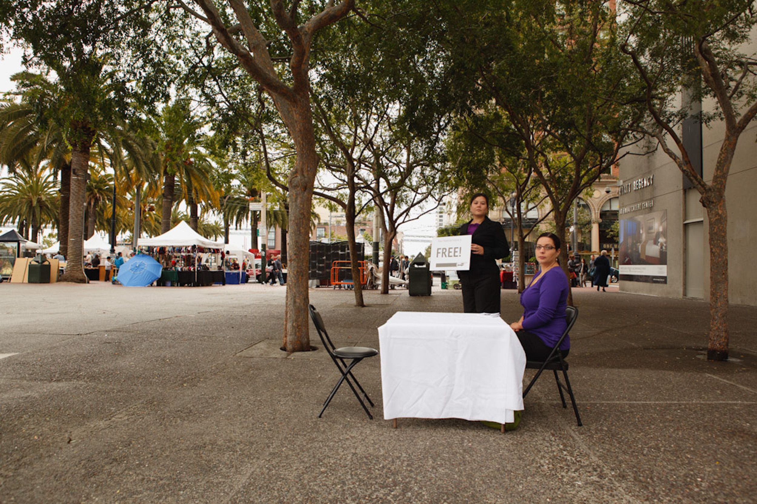 Tree-lined urban plaza with scattered seating areas and modern buildings in background.