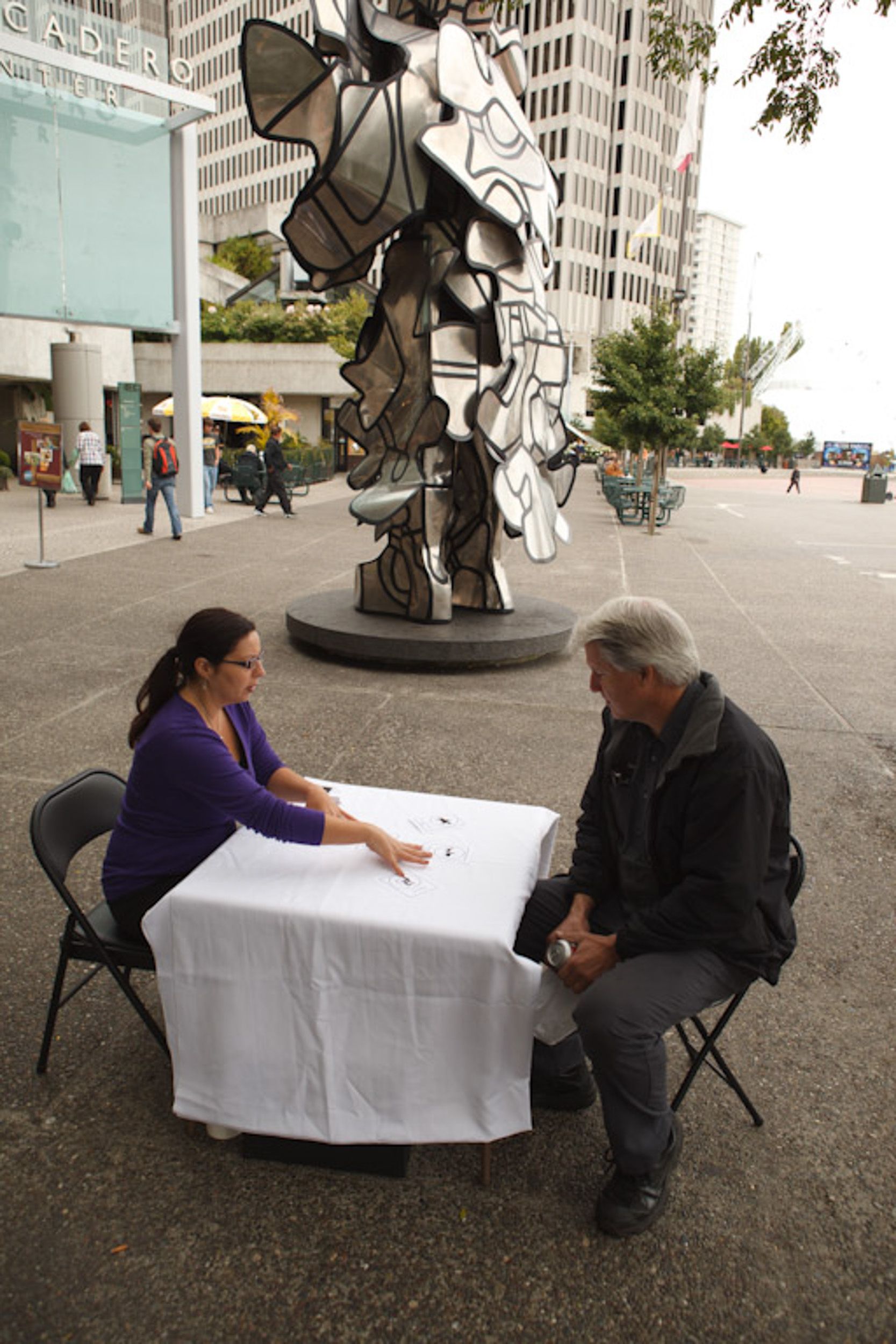 Two people sitting at a table with white tablecloth outdoors near urban buildings.
