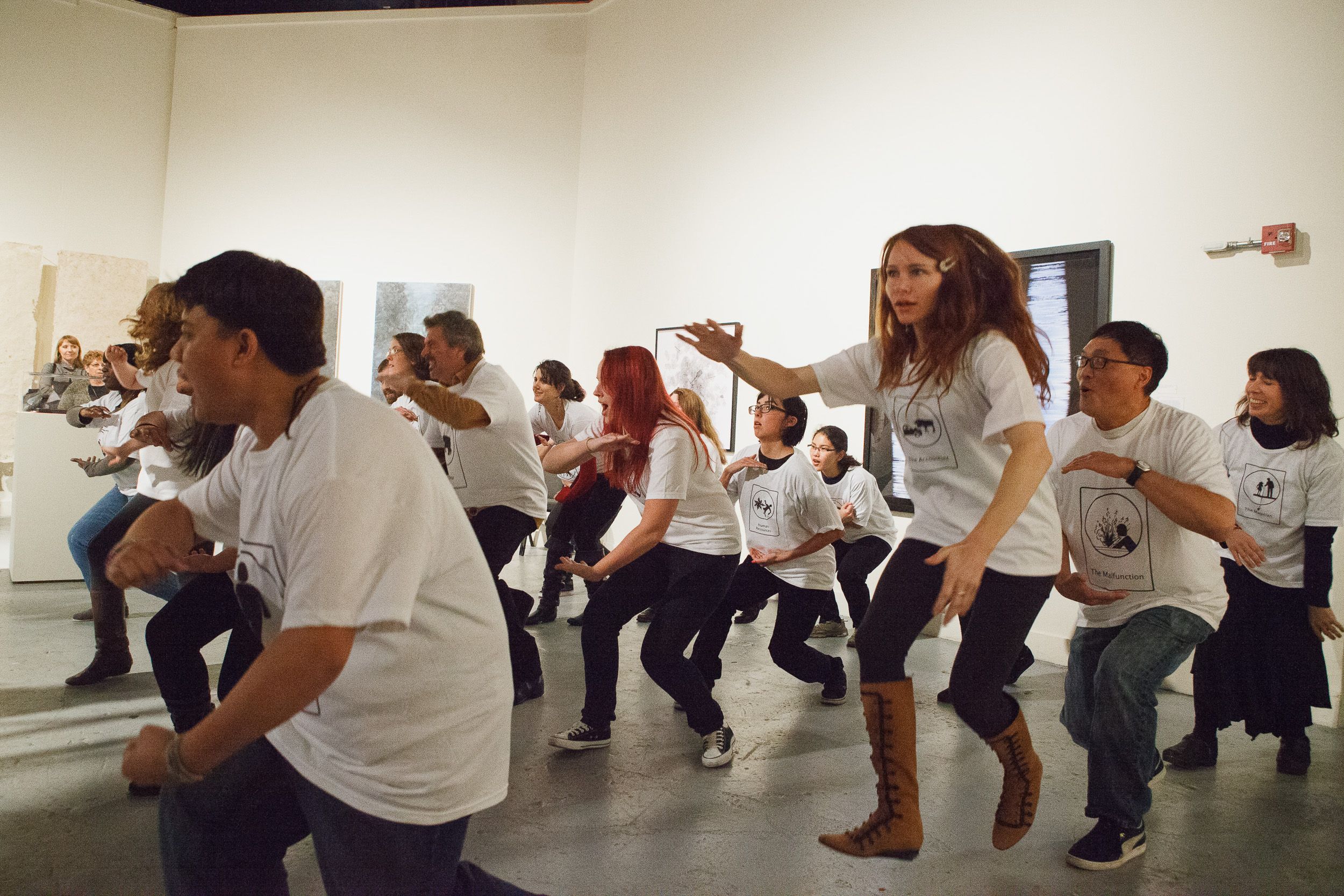 People in white shirts performing synchronized squatting movements in a gallery space.