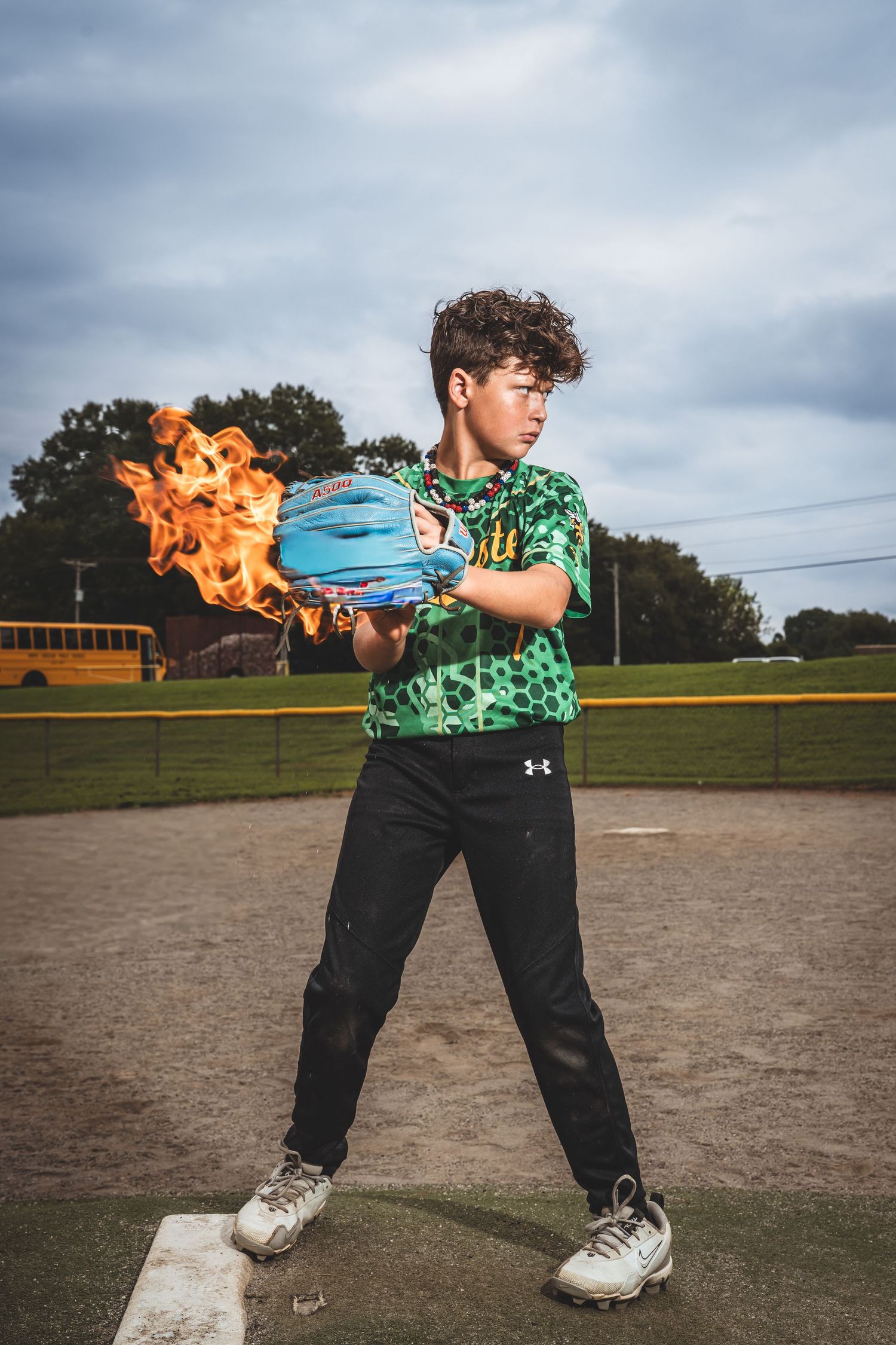 Person in a green patterned shirt performs fire breathing tricks on a baseball field at dusk.