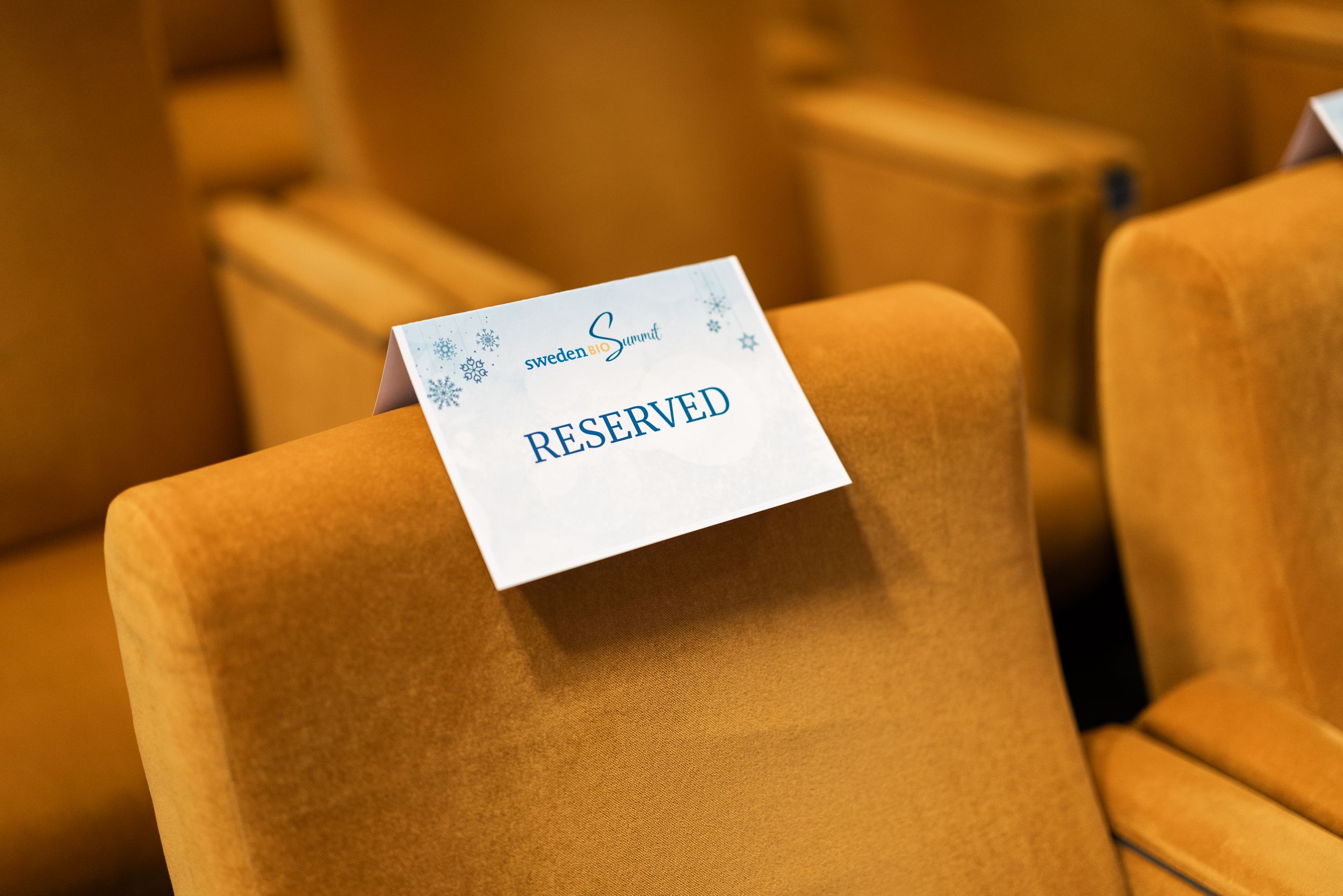 Rows of wooden chairs with white reserved signs placed on their seats in a warm-lit venue.