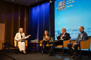 Speakers engage in discussion during a professional conference panel on stage with blue backdrop lighting.