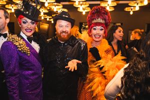 Colorful costumed performers in vibrant purple and orange outfits pose with someone in black at an indoor event with warm lighting.