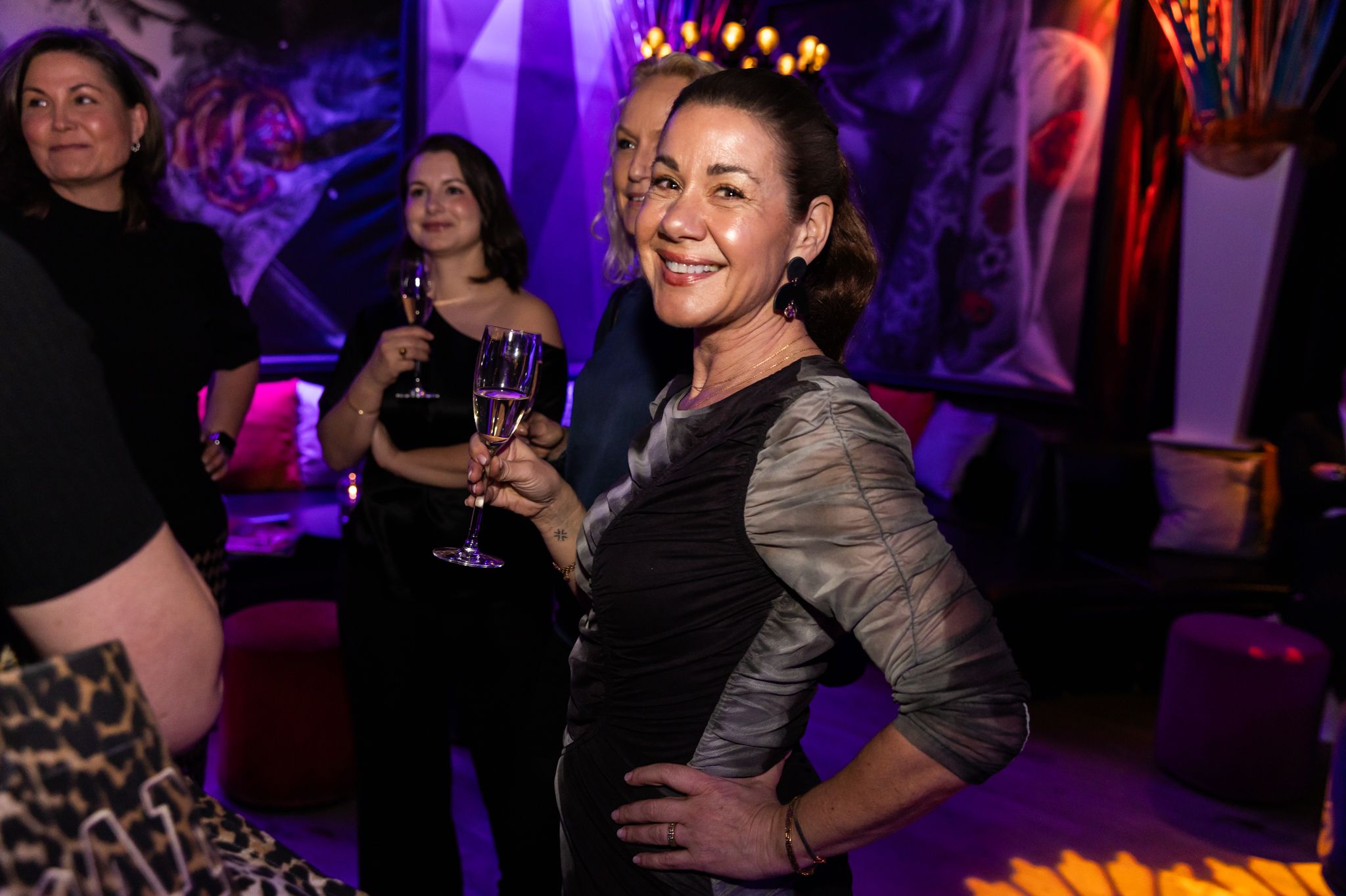 A person in a gray top smiles while holding a cocktail at a dimly lit party with purple lighting and chandeliers.