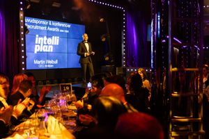 A speaker presents on stage at an evening business event with IntelliPlan displayed on screen while attendees dine at tables.