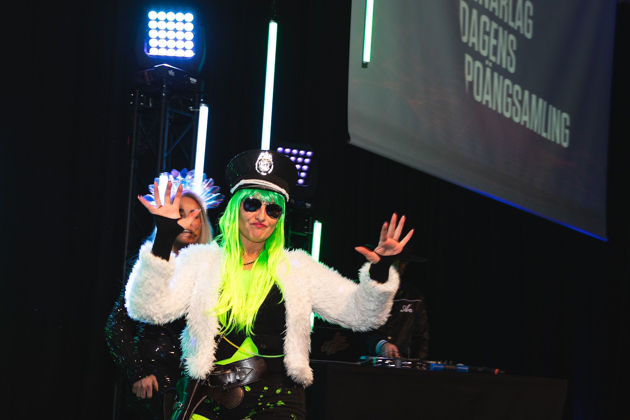 DJ wearing neon yellow jacket and white fur performs on stage with bright blue lights in background.
