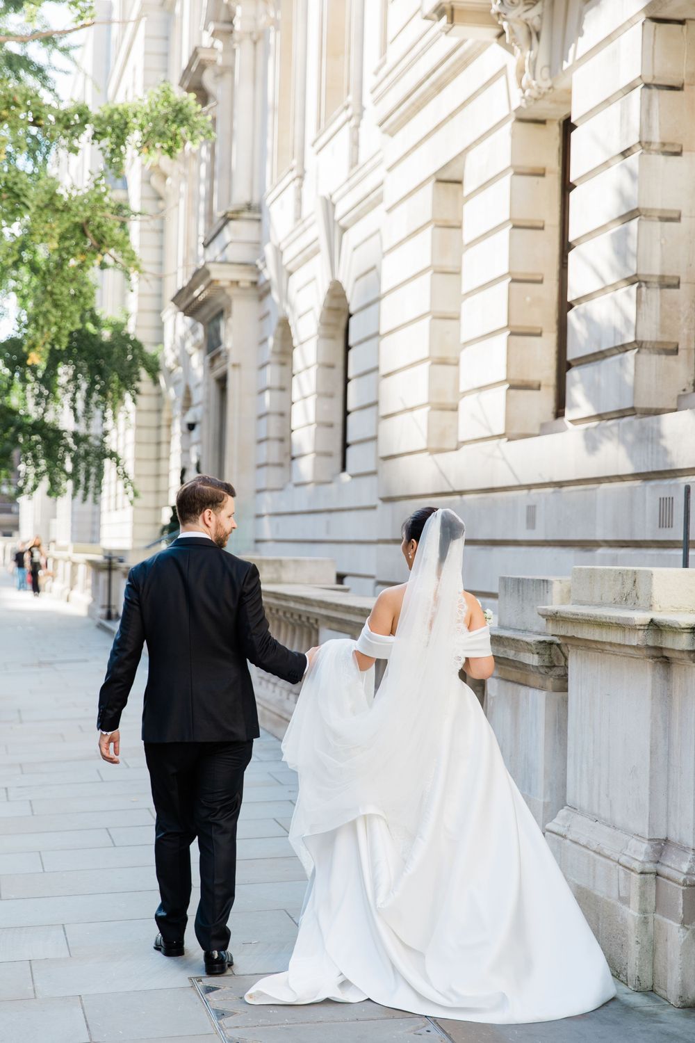 Newlyweds walking along grand stone building with flowing white wedding dress trailing behind at the Corinthia London by Maddie Waters