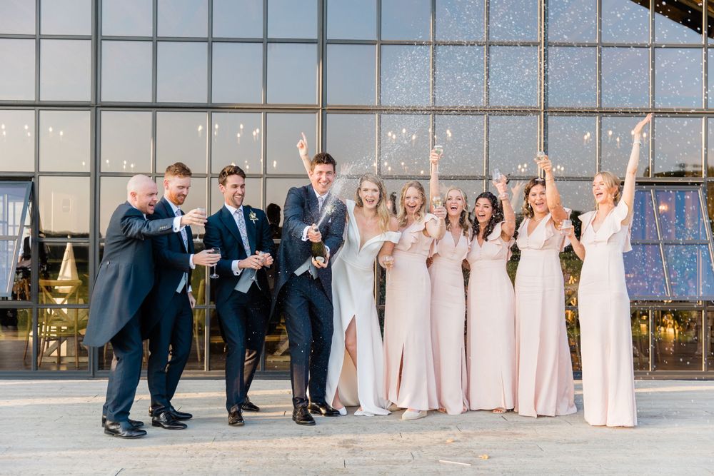 Wedding party celebrating with a champagne spray outside of The Barn at Botley Hill by Maddie Waters Surrey Photographer