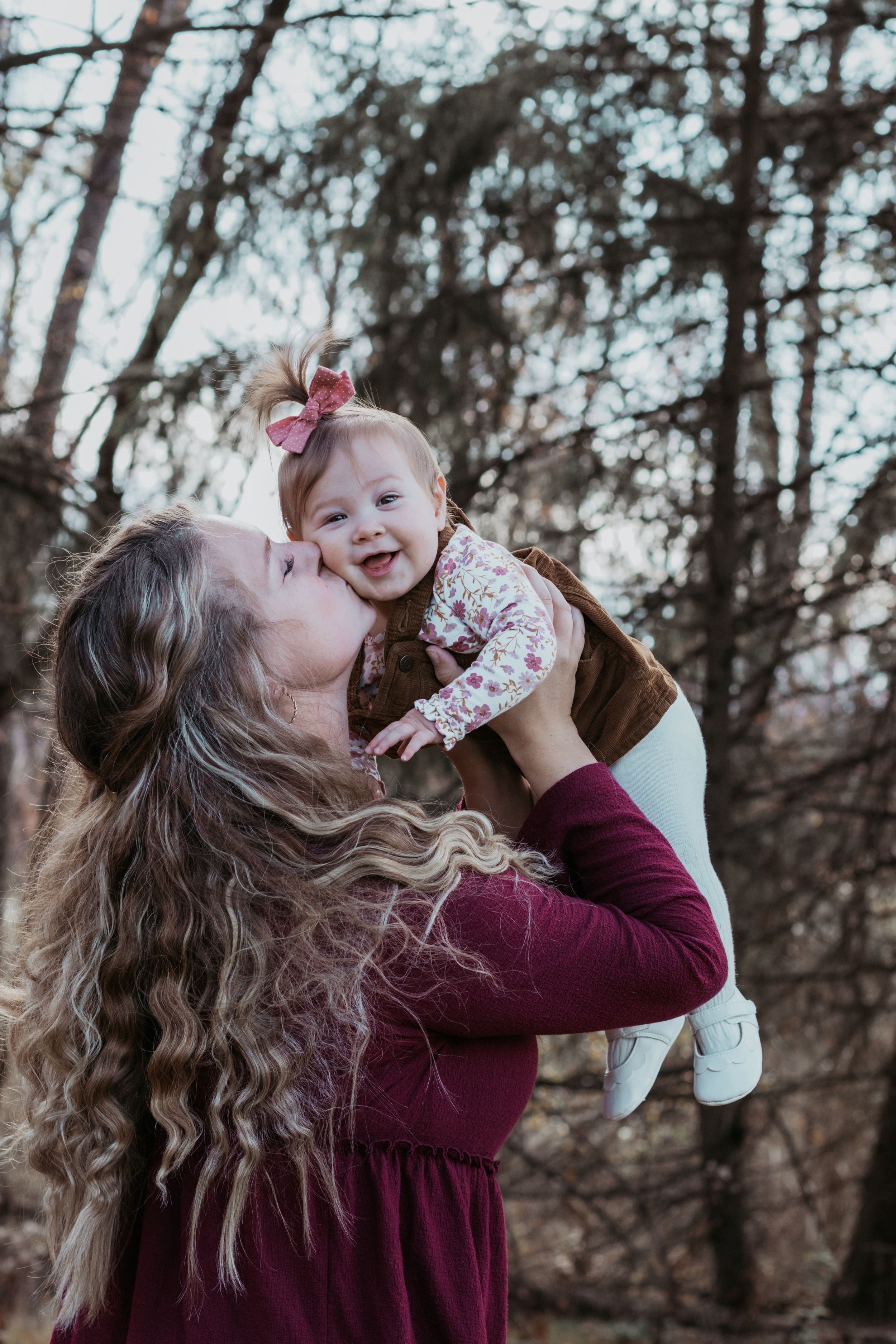 Mother lifting and kissing baby in playful moment against winter forest backdrop.