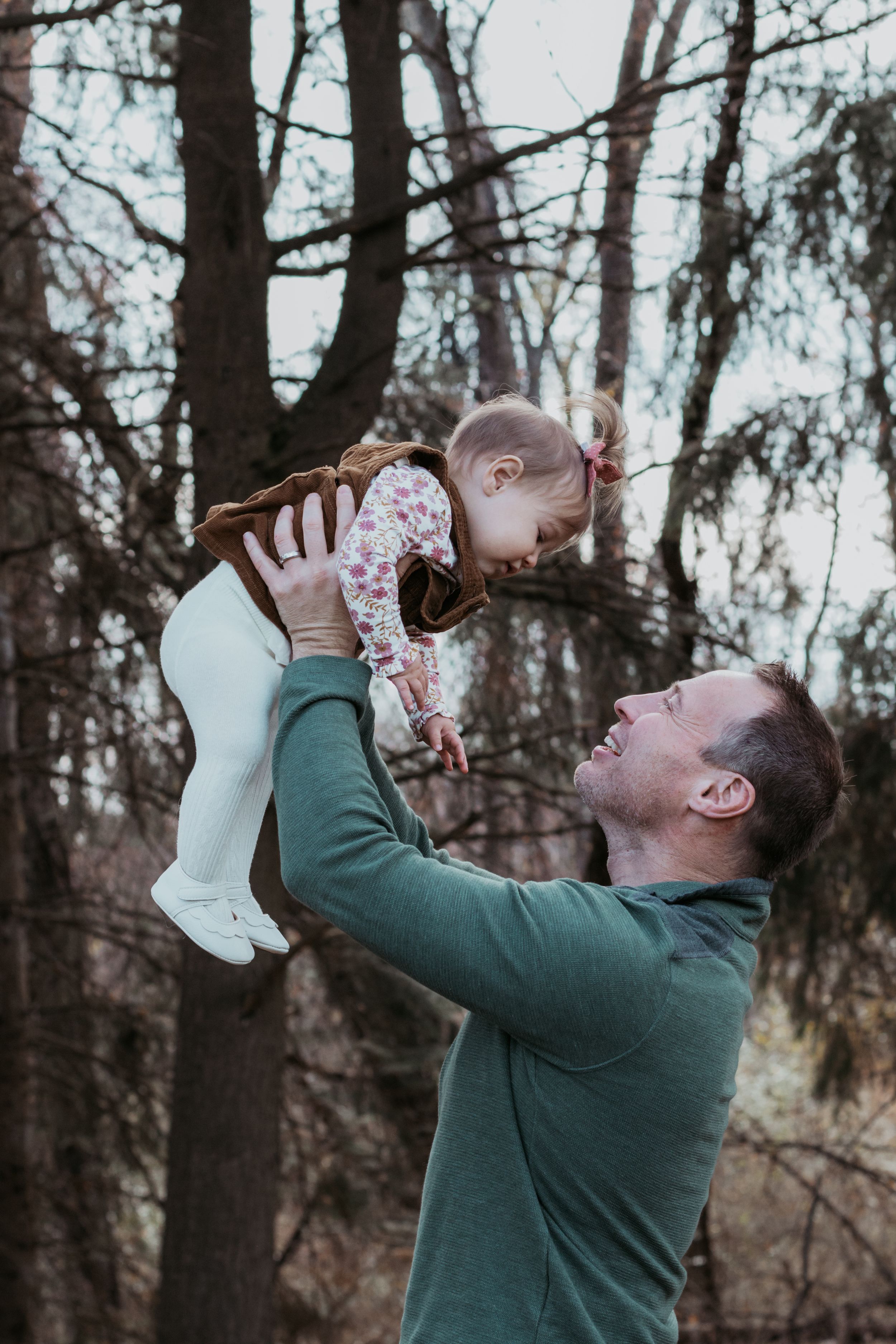 Parent lifting and playing with baby wearing floral outfit in wooded setting.