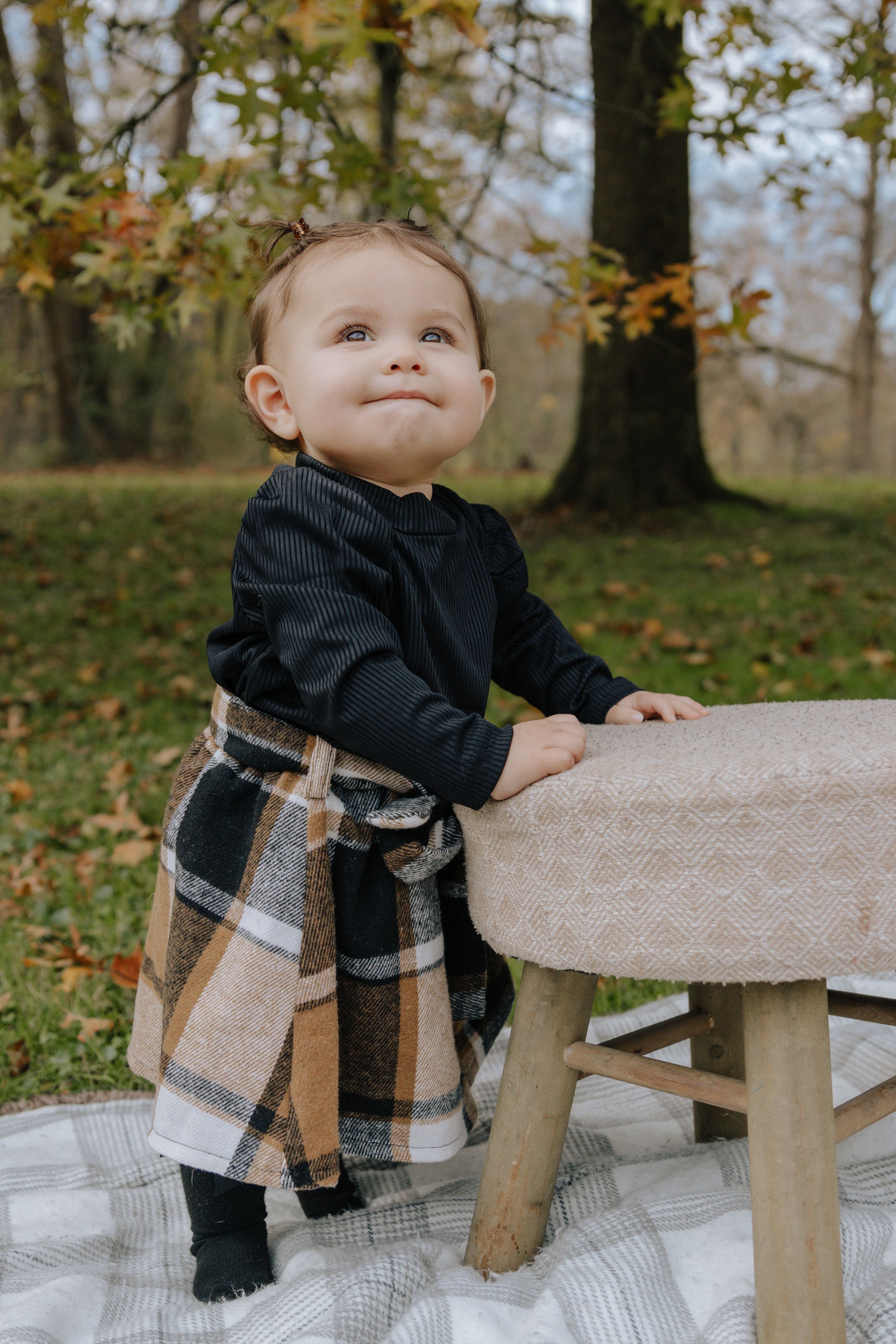 Small child in black sweater and plaid pants leans against rustic bench in autumn forest setting.