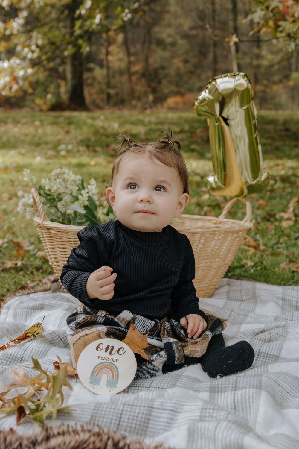 First birthday photoshoot setup with child on blanket next to wicker basket and gold number one balloon.