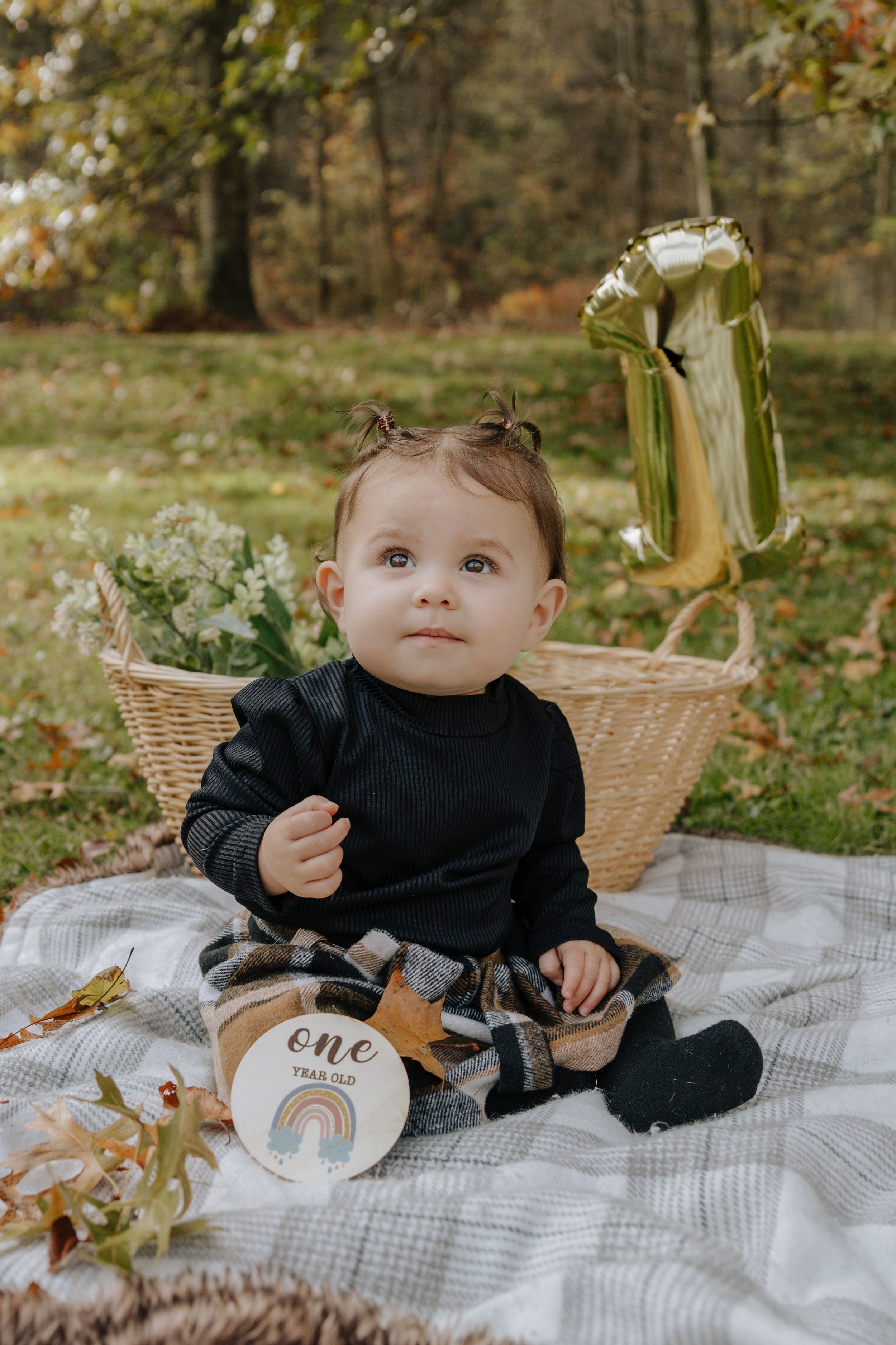First birthday photoshoot setup with child on blanket next to wicker basket and gold number one balloon.