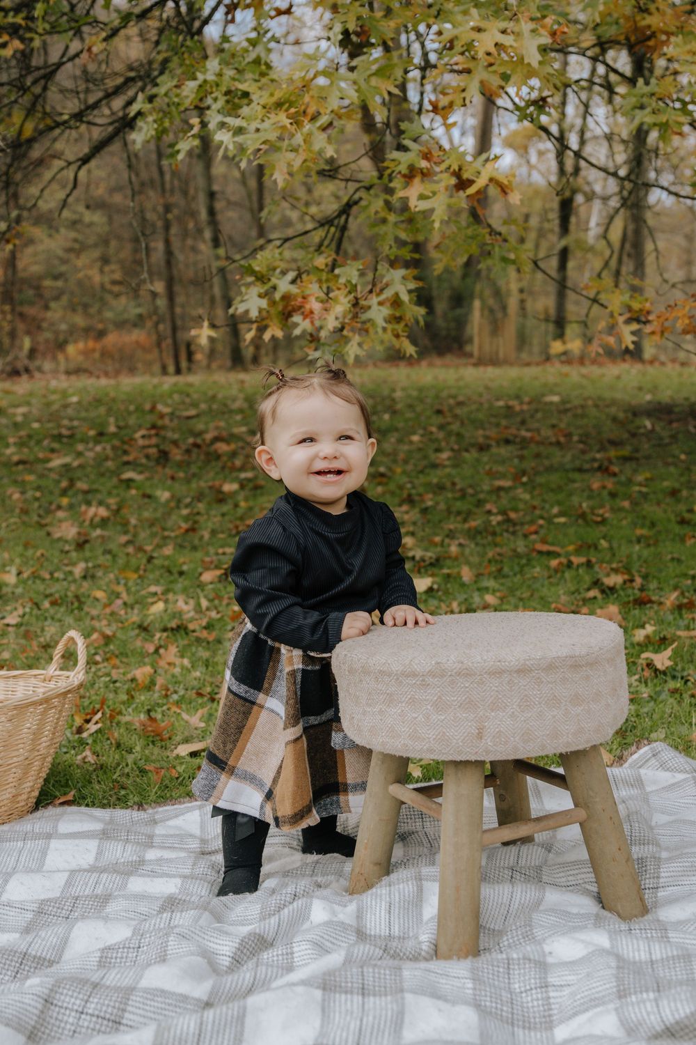 Small child in dark outfit stands beside wooden stool on picnic blanket in autumn setting.