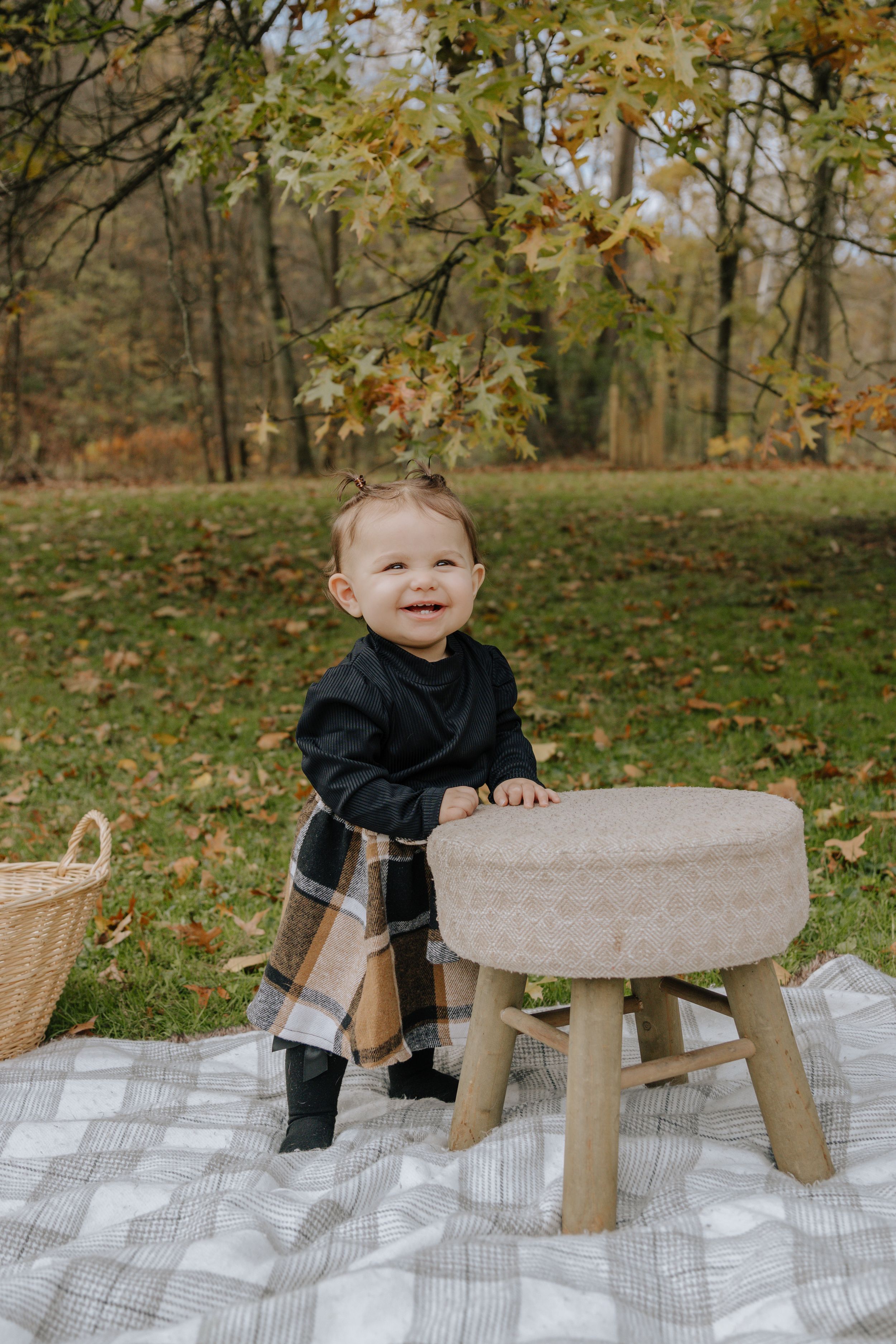 Small child in dark outfit stands beside wooden stool on picnic blanket in autumn setting.