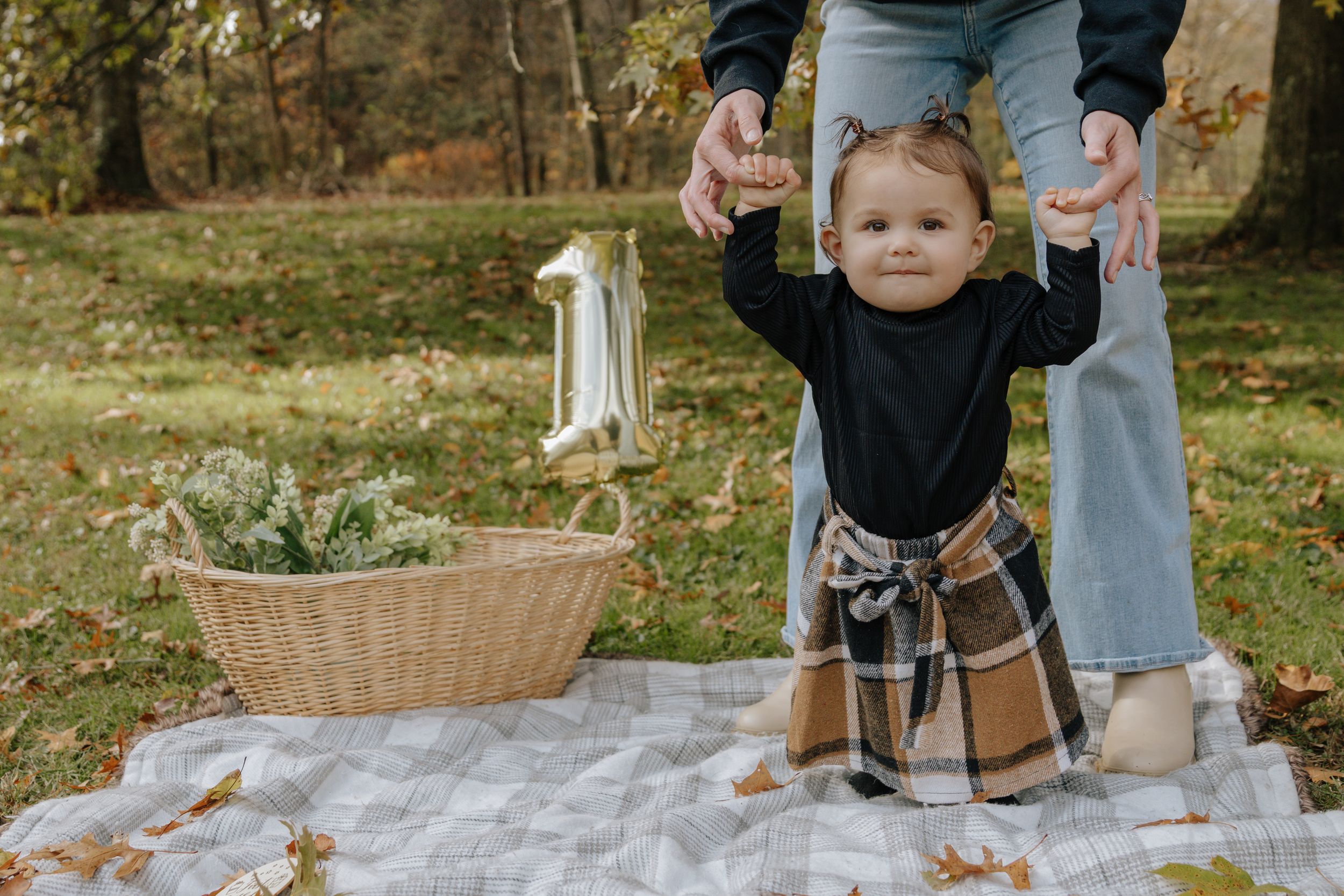 Child standing on picnic blanket reaching up while supported in autumn outdoor setting.