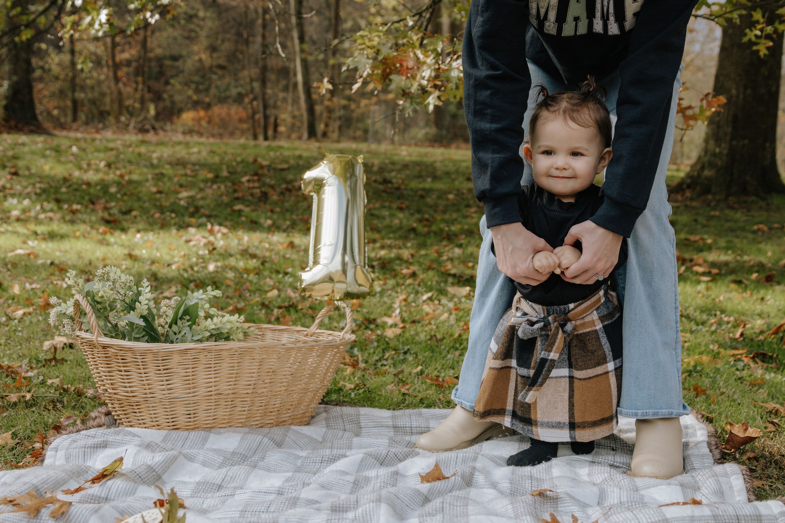 Child in plaid pants and black top standing on blanket with support in wooded setting.