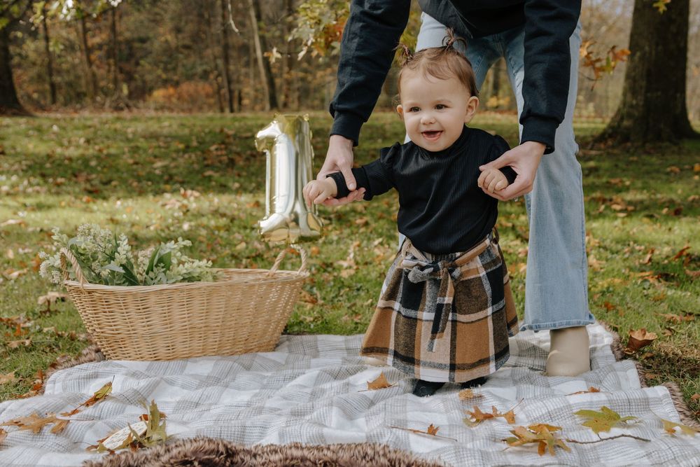 Child taking steps on picnic blanket with assistance during fall outdoor photoshoot.