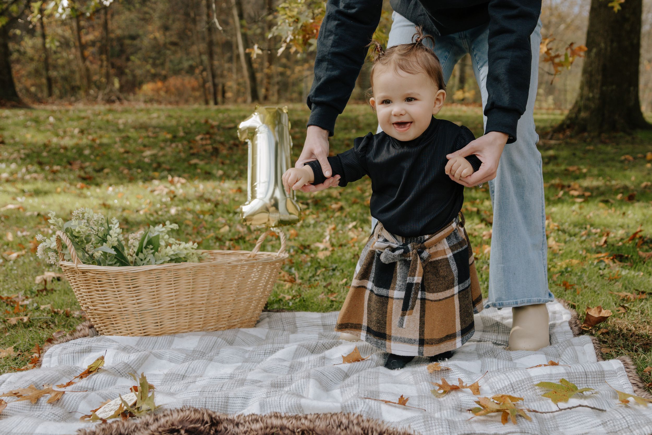 Child taking steps on picnic blanket with assistance during fall outdoor photoshoot.