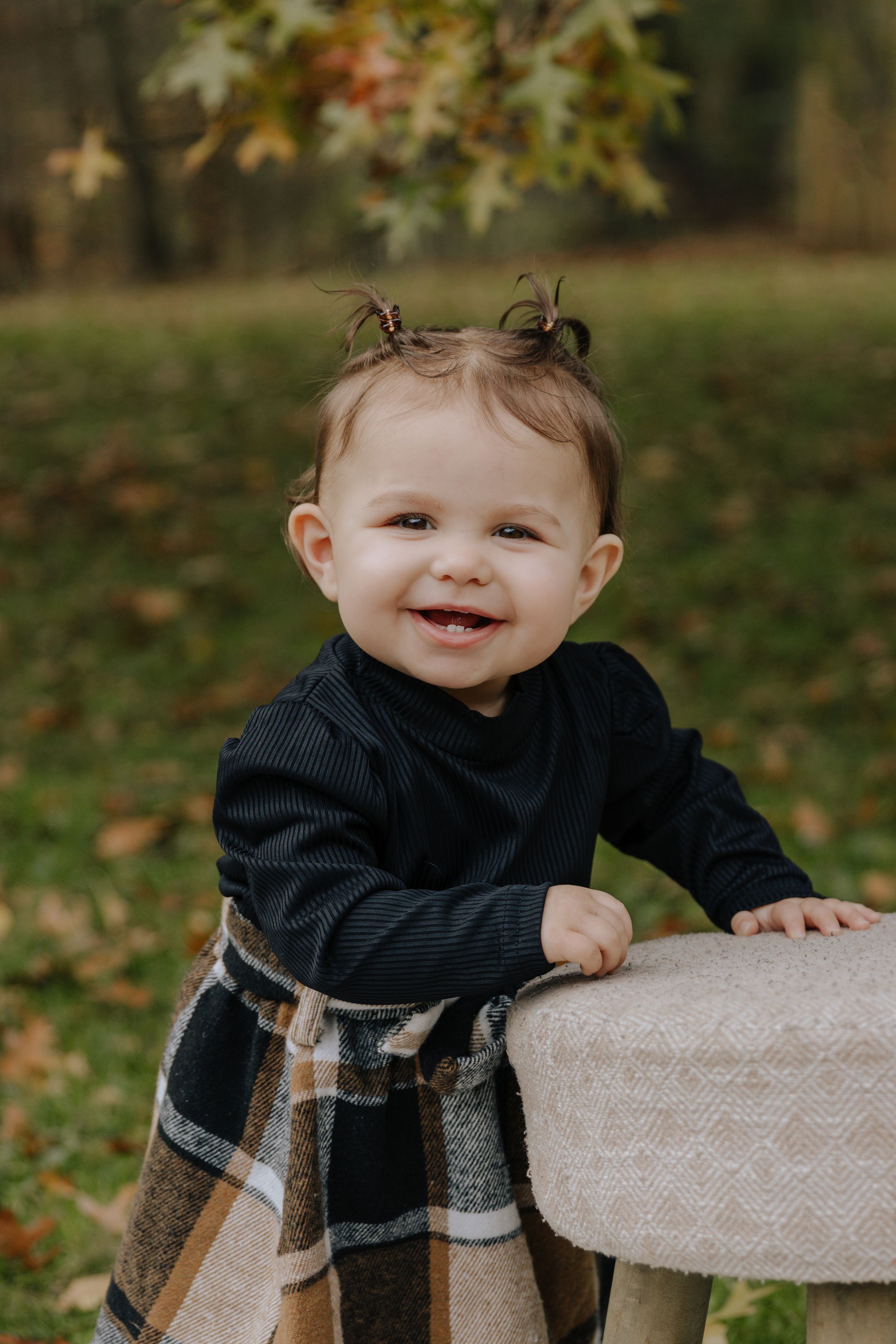 Toddler in dark outfit and plaid skirt leans on wooden stool outdoors.