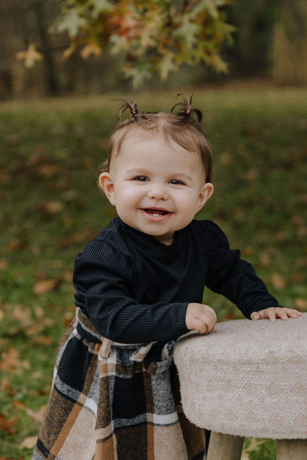 Toddler in dark outfit and plaid skirt leans on wooden stool outdoors.