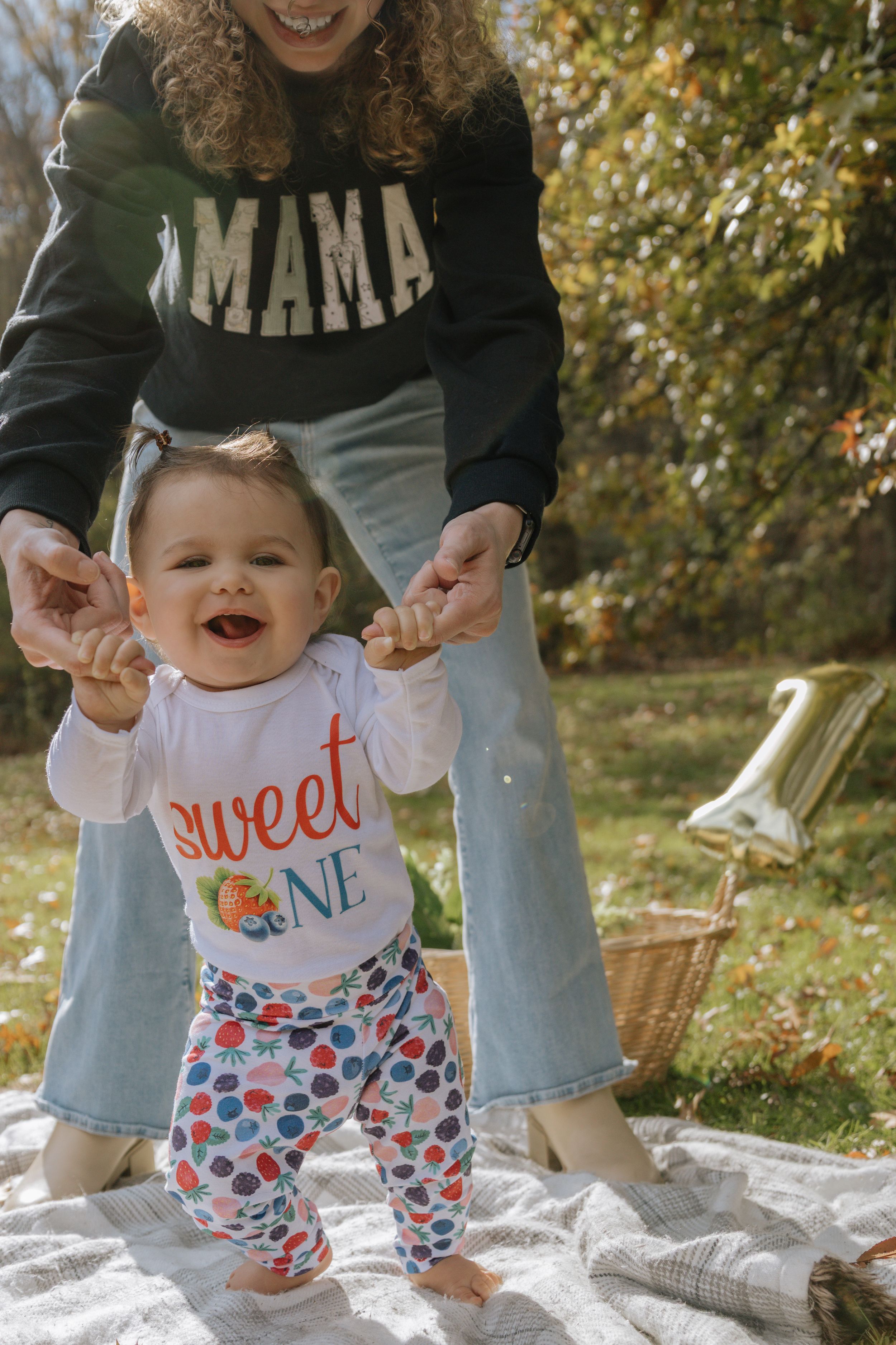 A baby in a 'sweet one' shirt and floral pants stands on a blanket while being supported by someone in a mama sweatshirt.