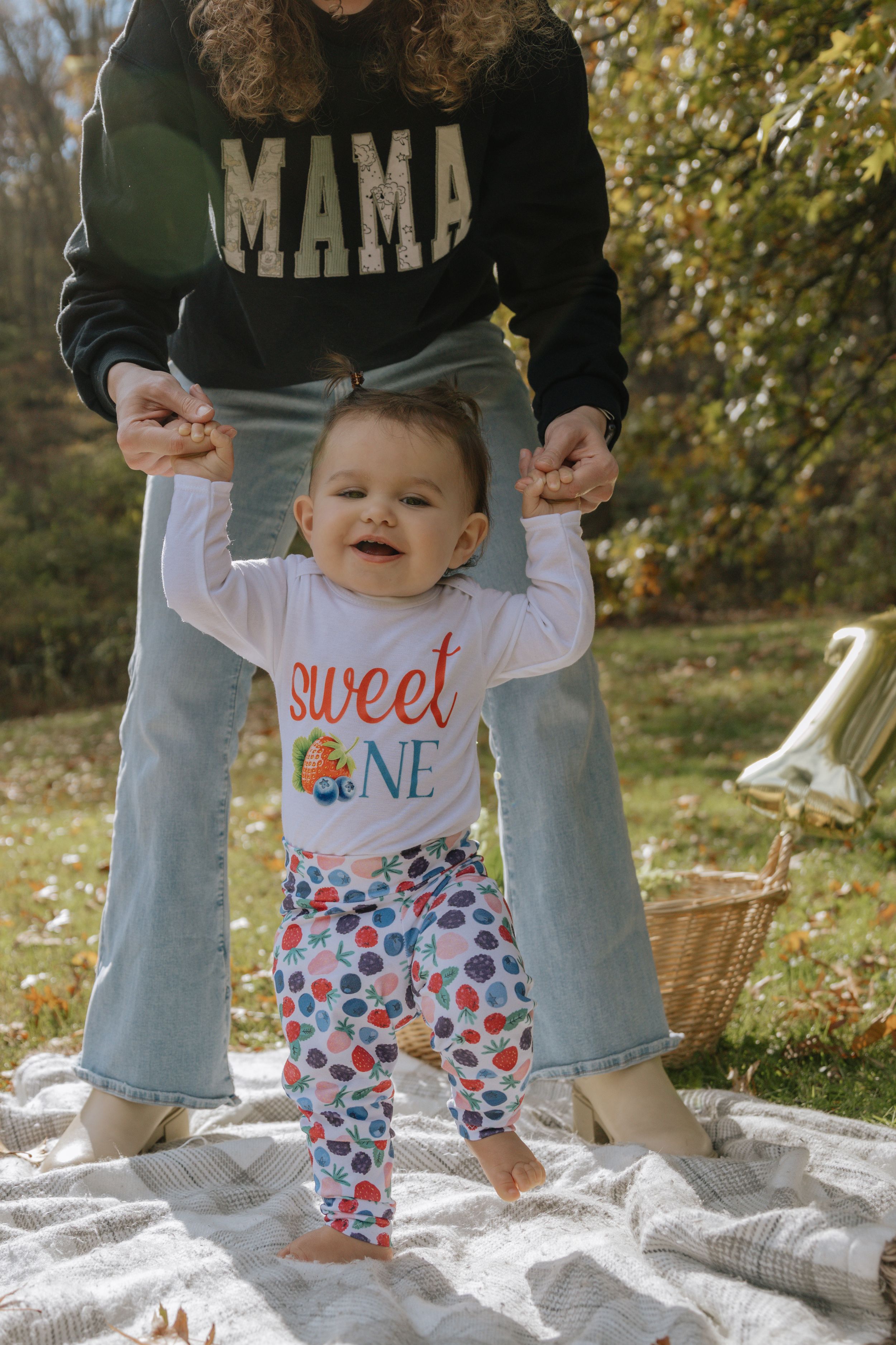 A smiling baby wearing a 'sweet one' shirt stands on a white blanket outdoors during autumn.