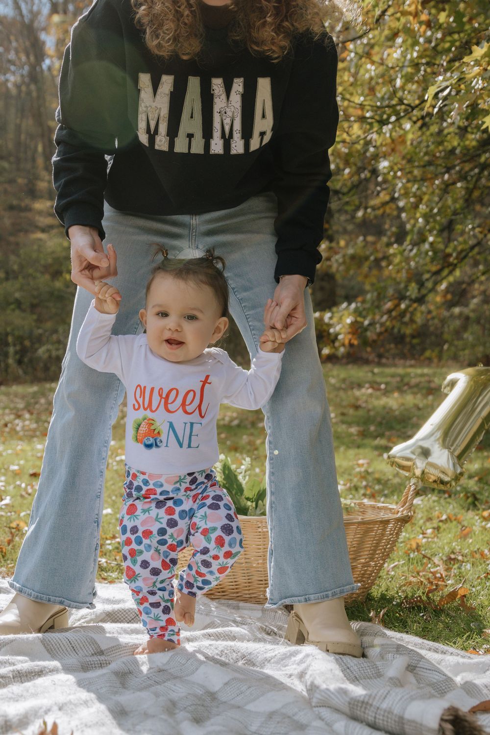 A baby in colorful pants takes first steps on a picnic blanket while being supported.