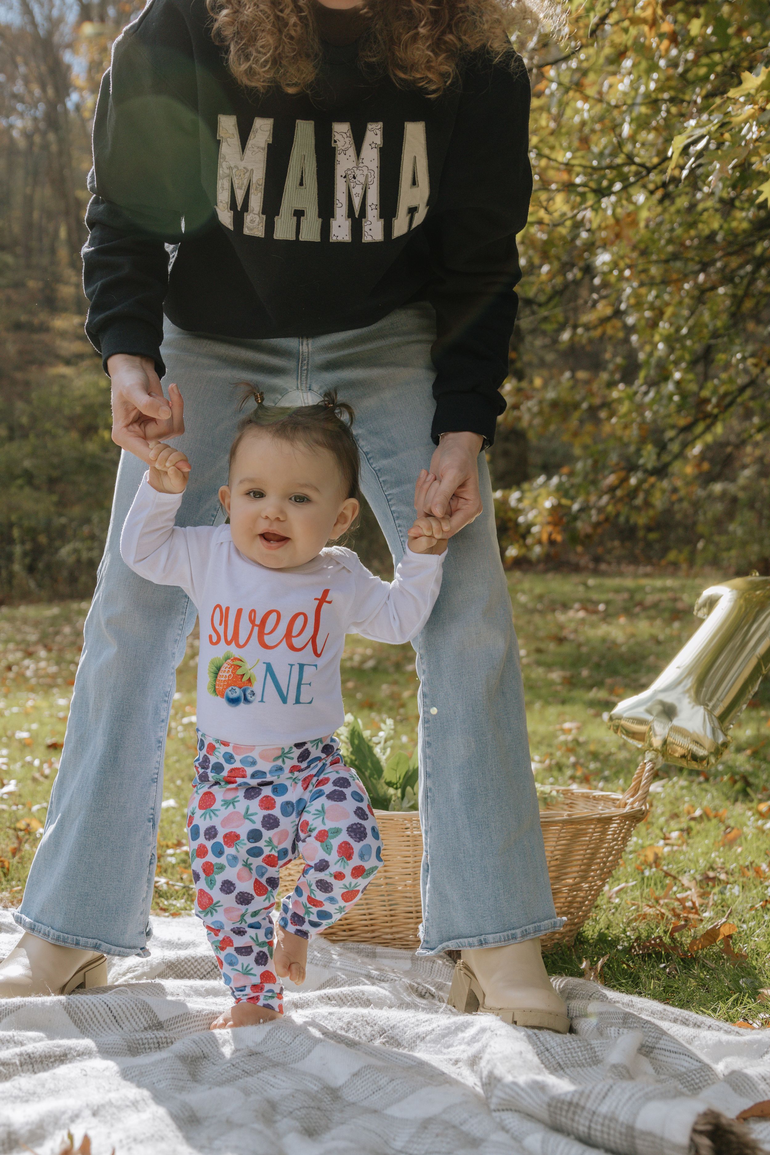 A baby in colorful pants takes first steps on a picnic blanket while being supported.