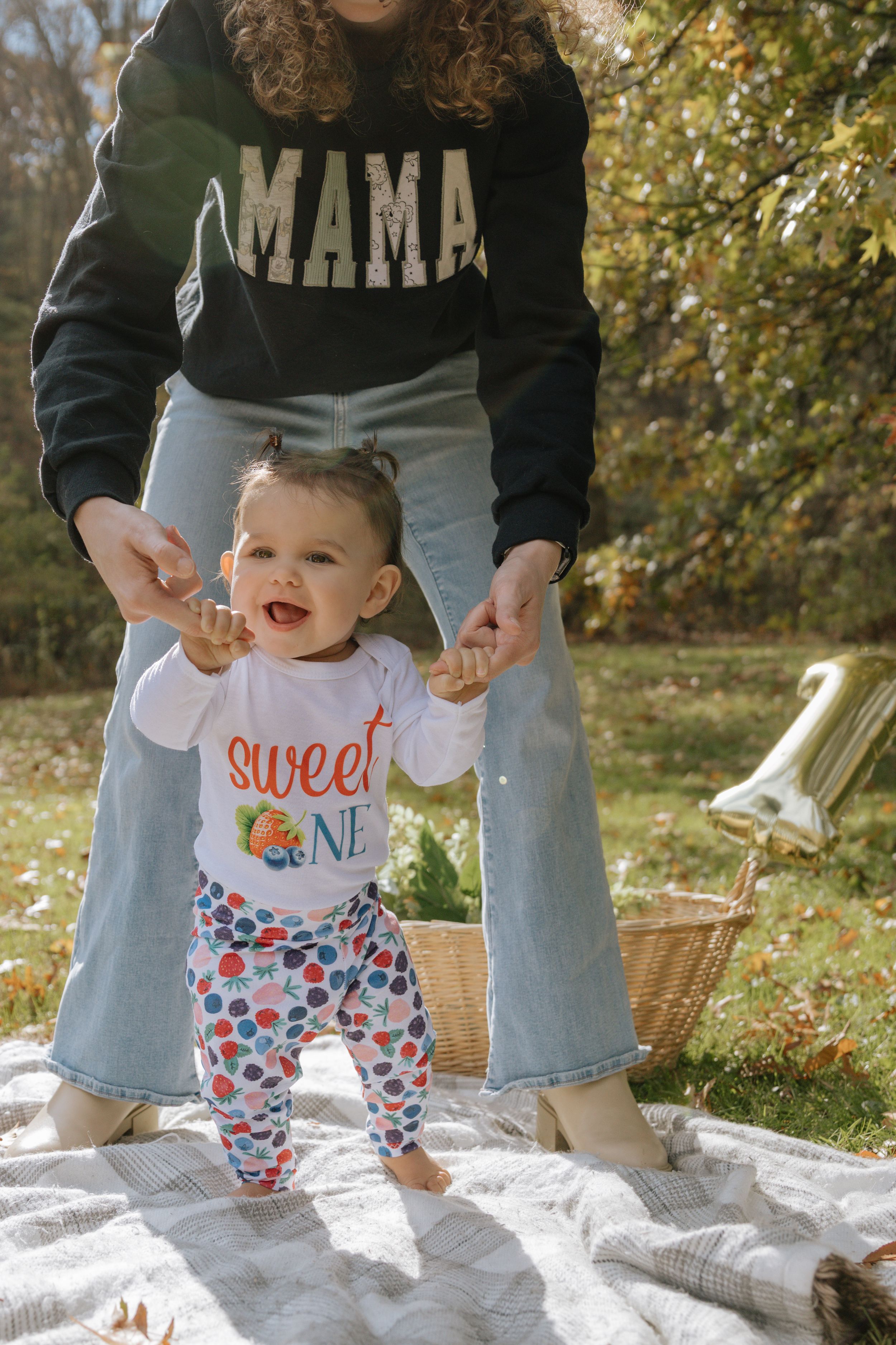 A joyful baby practices standing on a blanket outdoors while wearing a festive outfit.