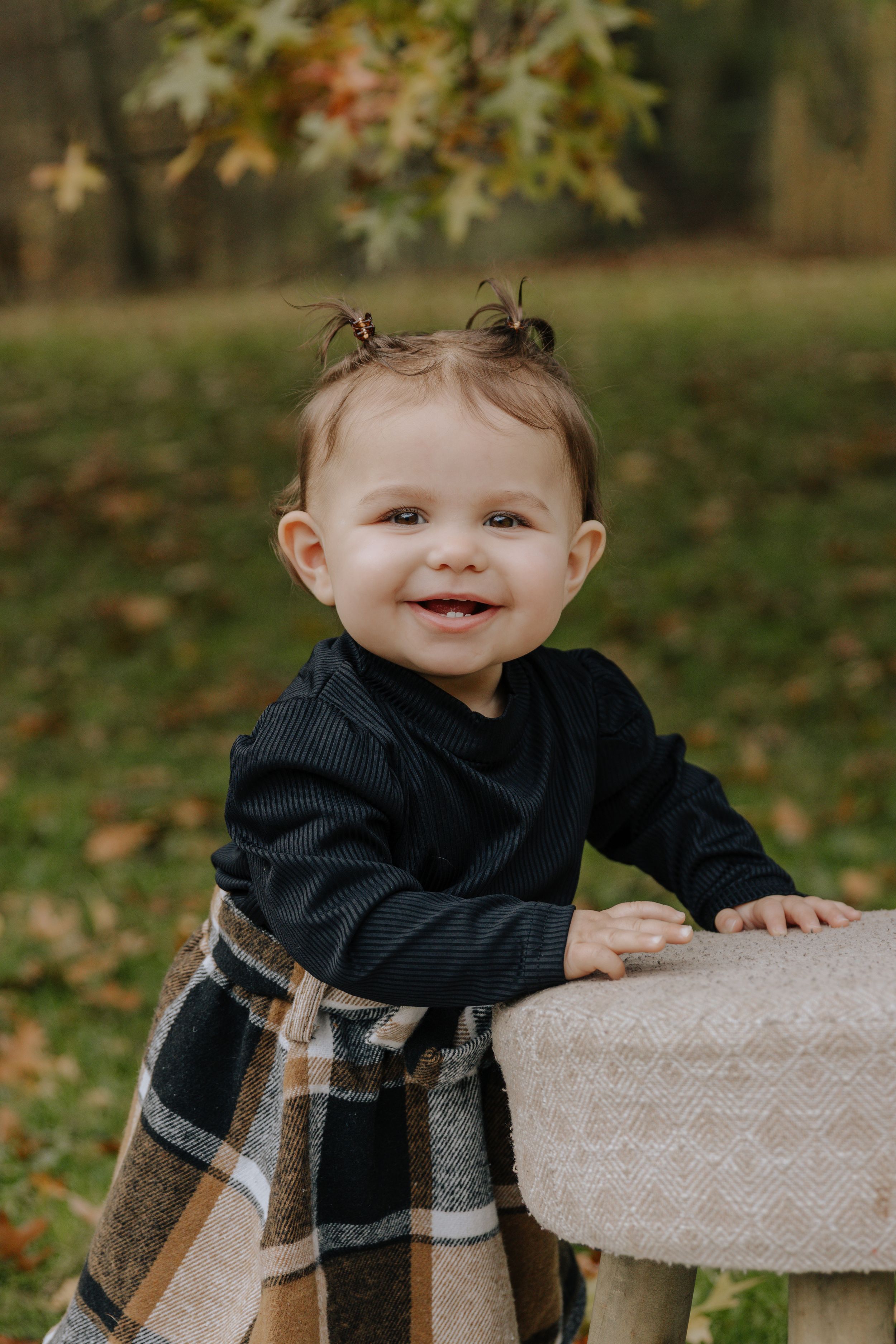 Happy toddler with pigtails wearing dark outfit leans on wooden stool in autumn setting.