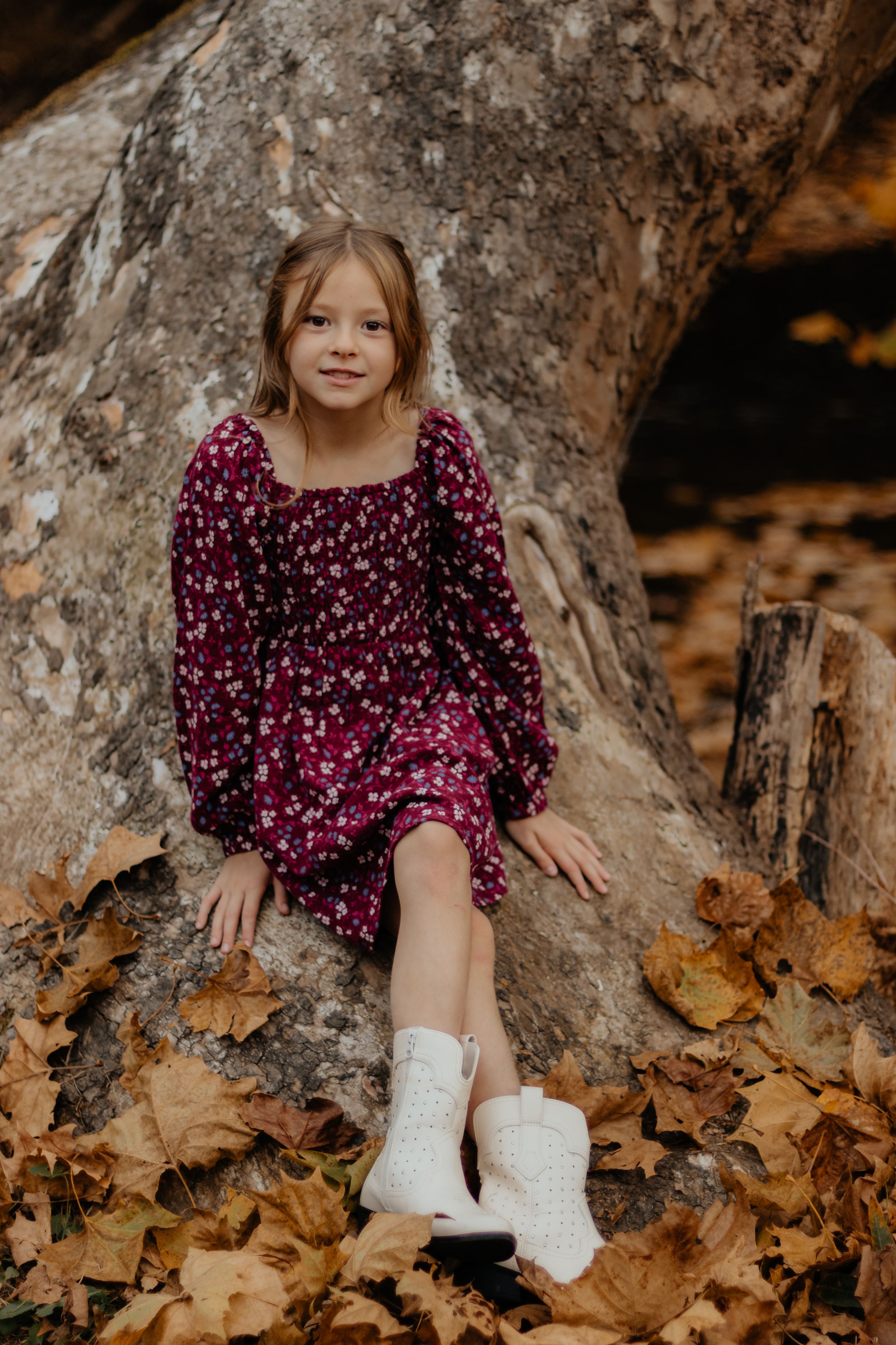 Child in burgundy polka dot dress sits on rock surrounded by fallen autumn leaves.
