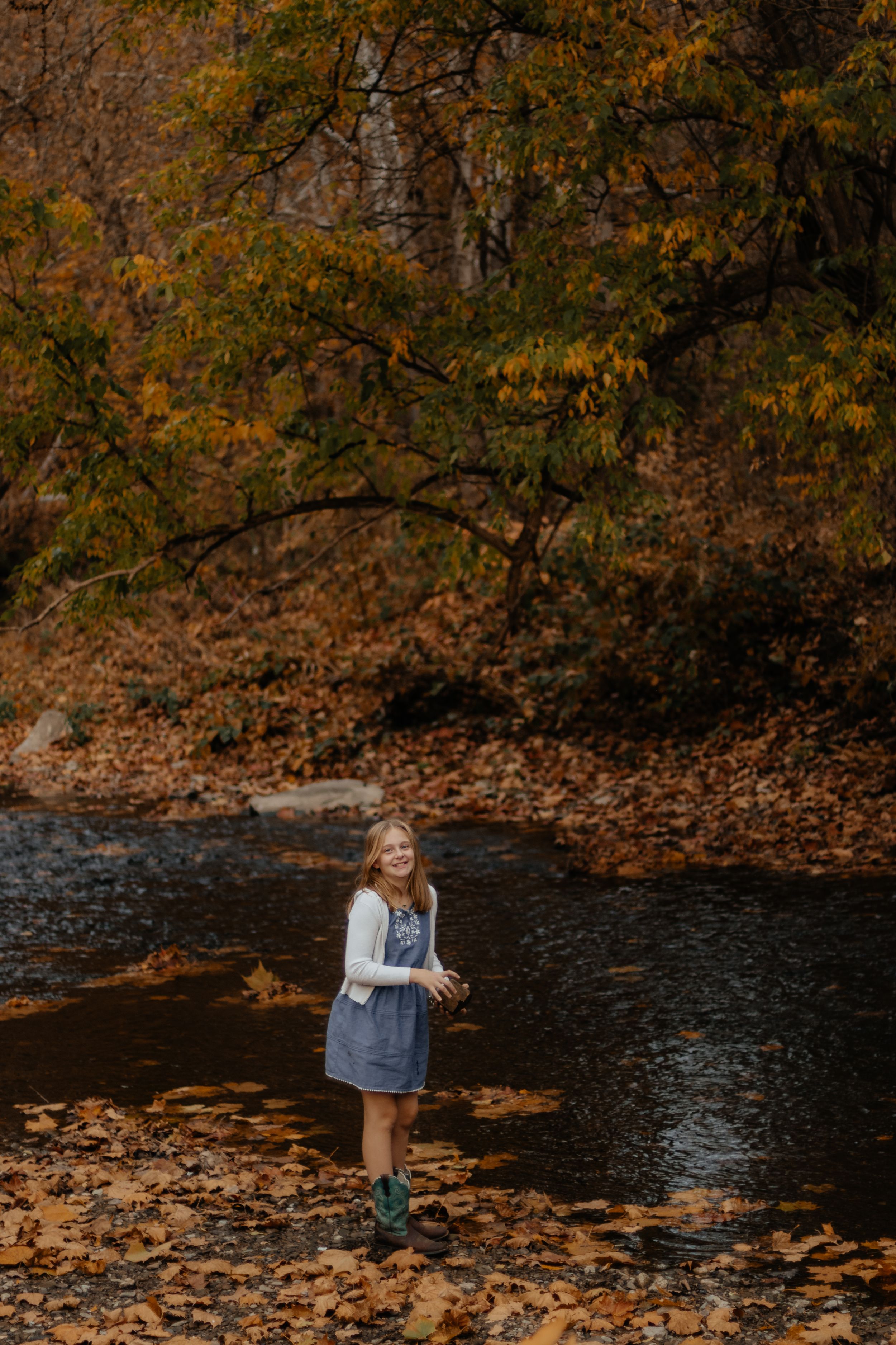 Child in denim dress explores near water's edge surrounded by fall foliage.