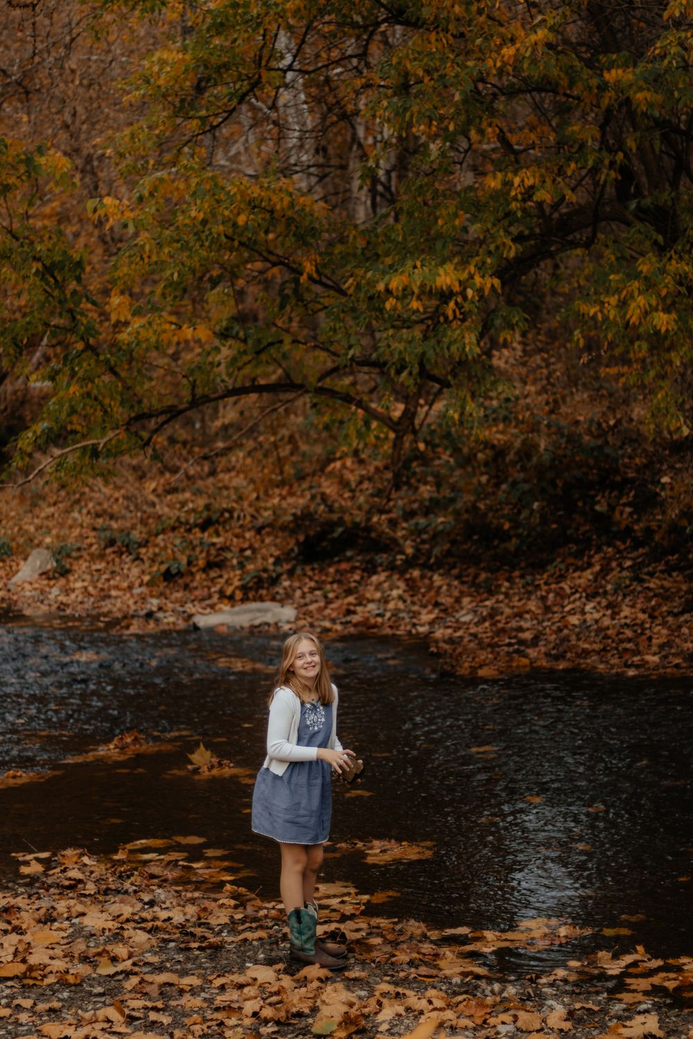 Child in denim dress explores near water's edge surrounded by fall foliage.