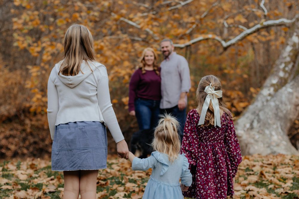 Family walking away from camera through autumn woods with fallen leaves on ground.
