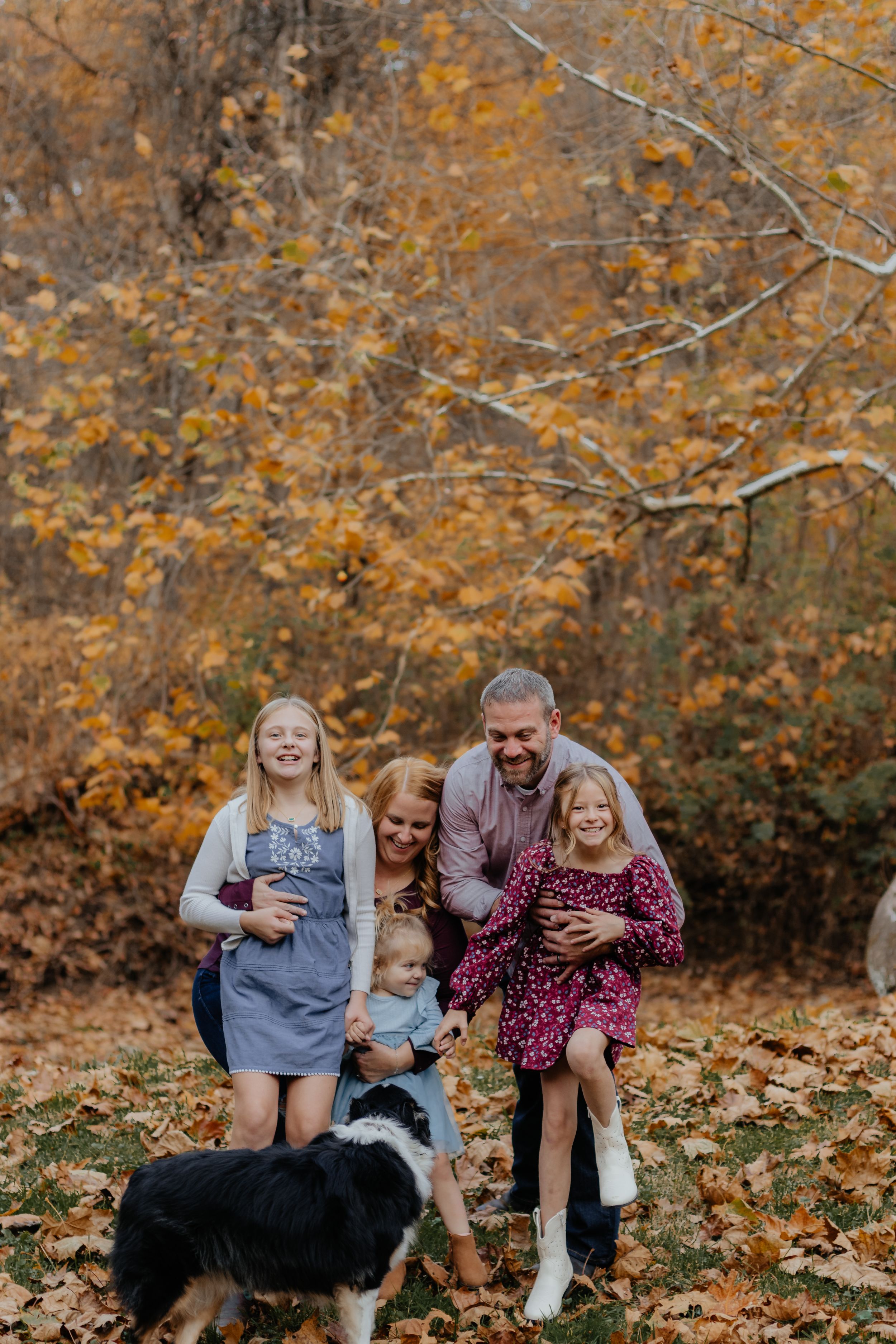 Family group plays and laughs together on ground covered with autumn leaves.