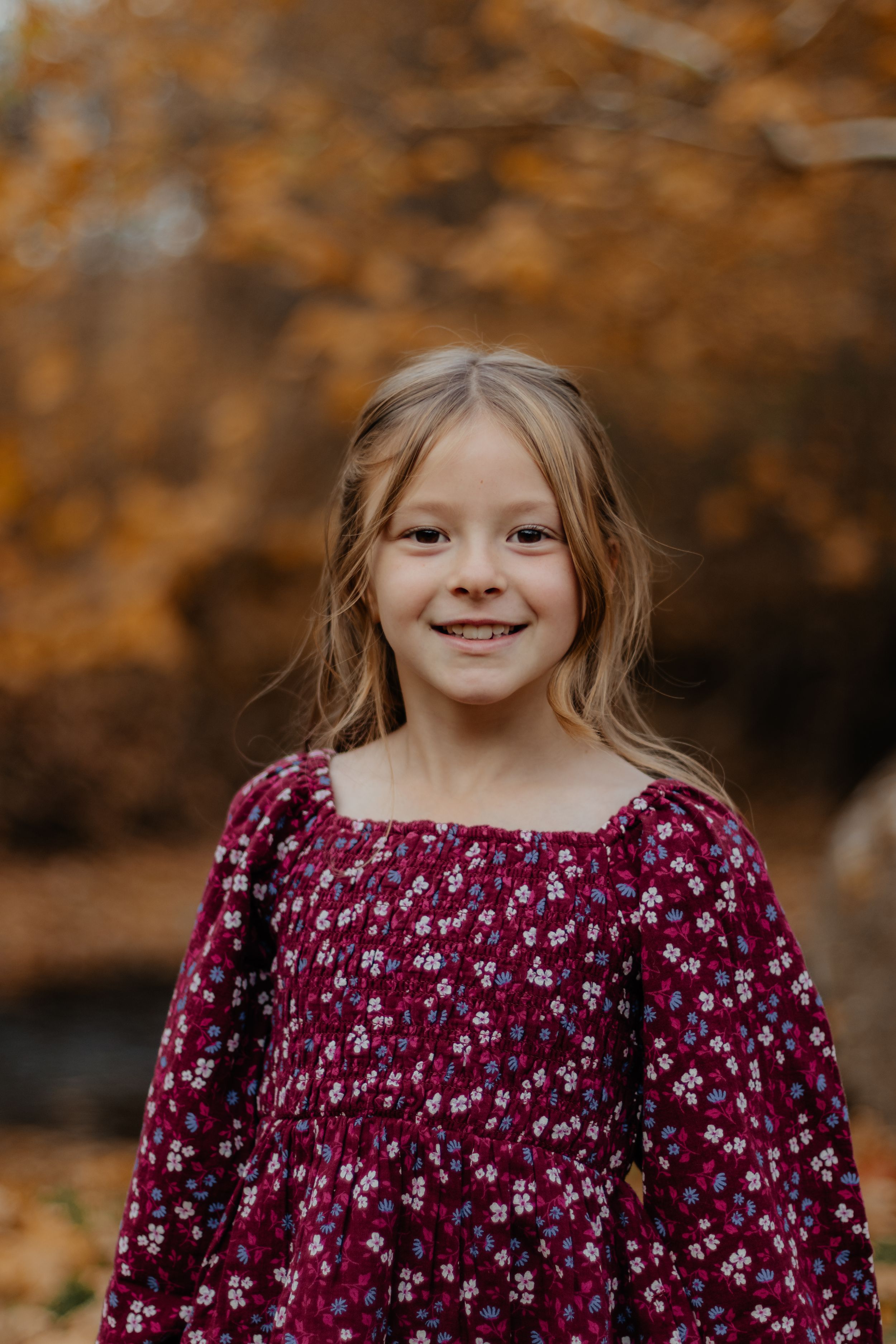Portrait against autumn foliage background showing burgundy floral dress and warm lighting.