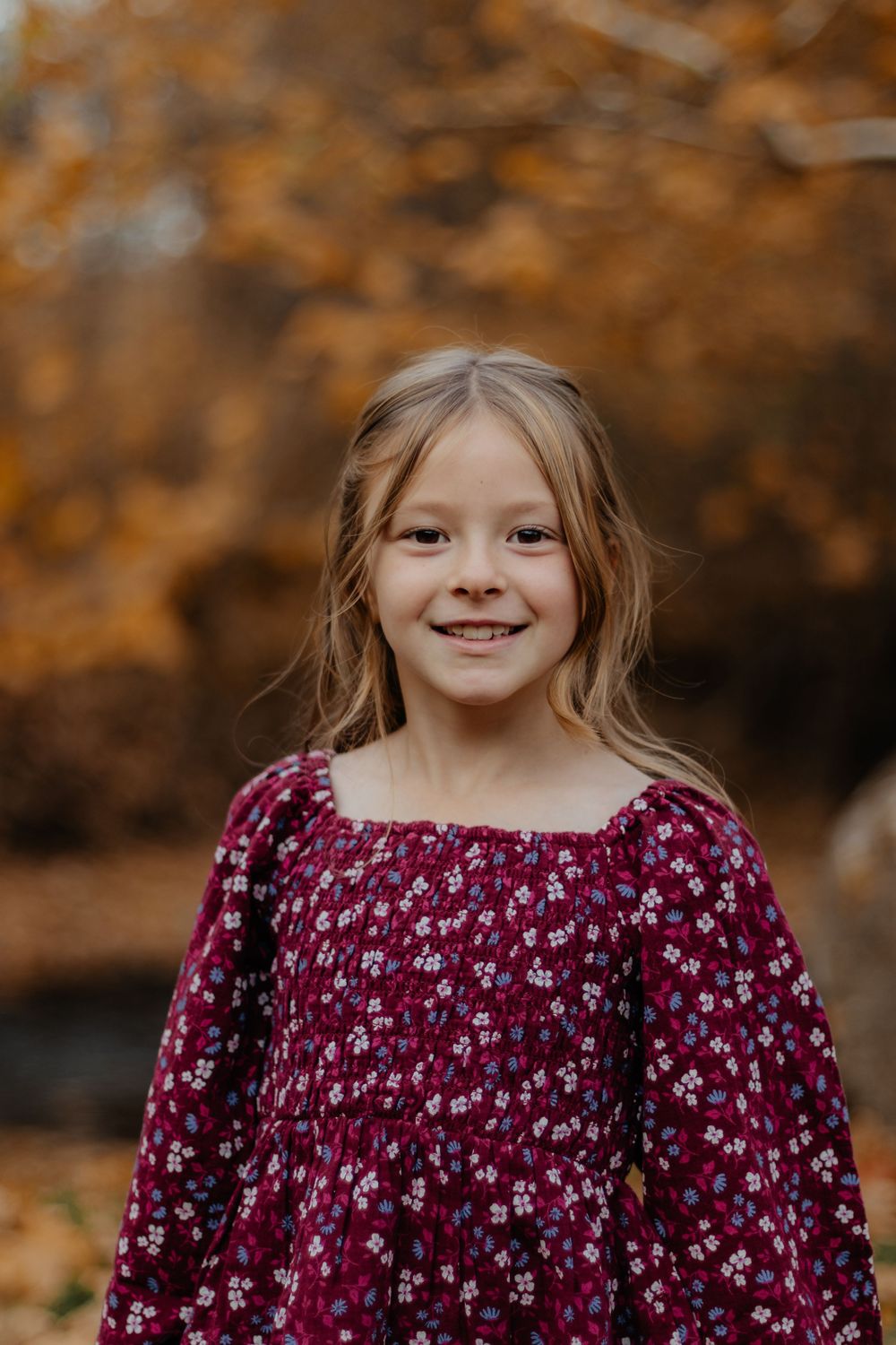 Portrait against autumn foliage background showing burgundy floral dress and warm lighting.