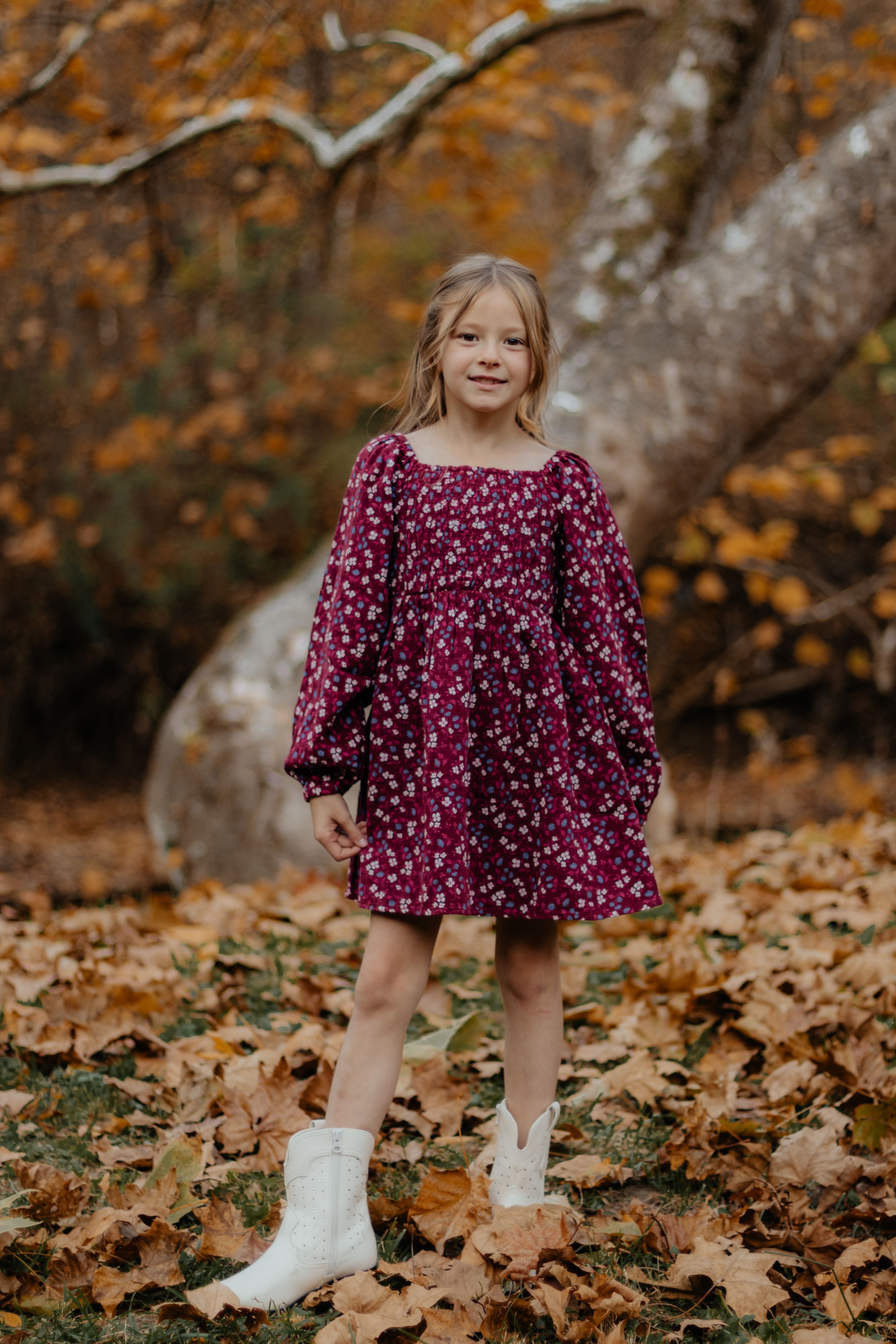 Standing pose in burgundy floral dress with white boots against fall forest backdrop.