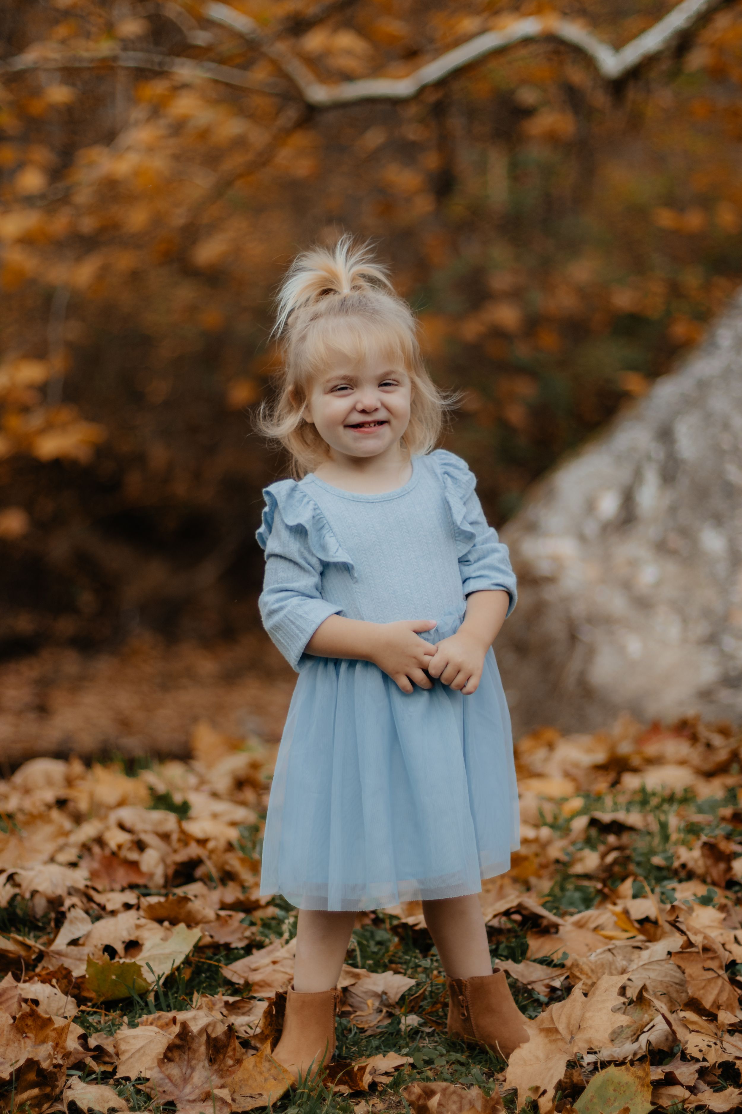 Cheerful pose in powder blue dress standing among fallen autumn leaves.