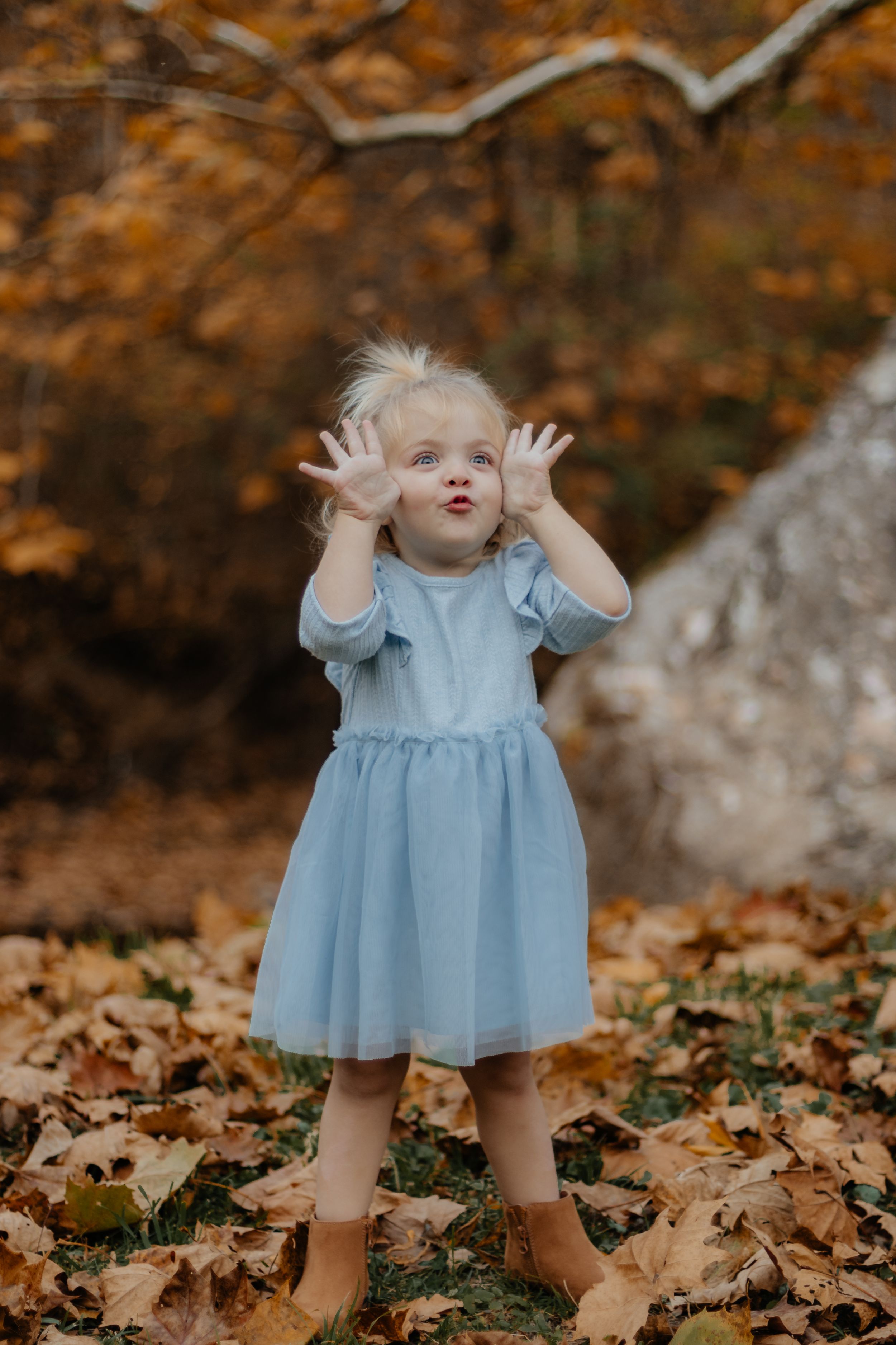 Expressive pose in light blue dress with hands raised against autumn background.