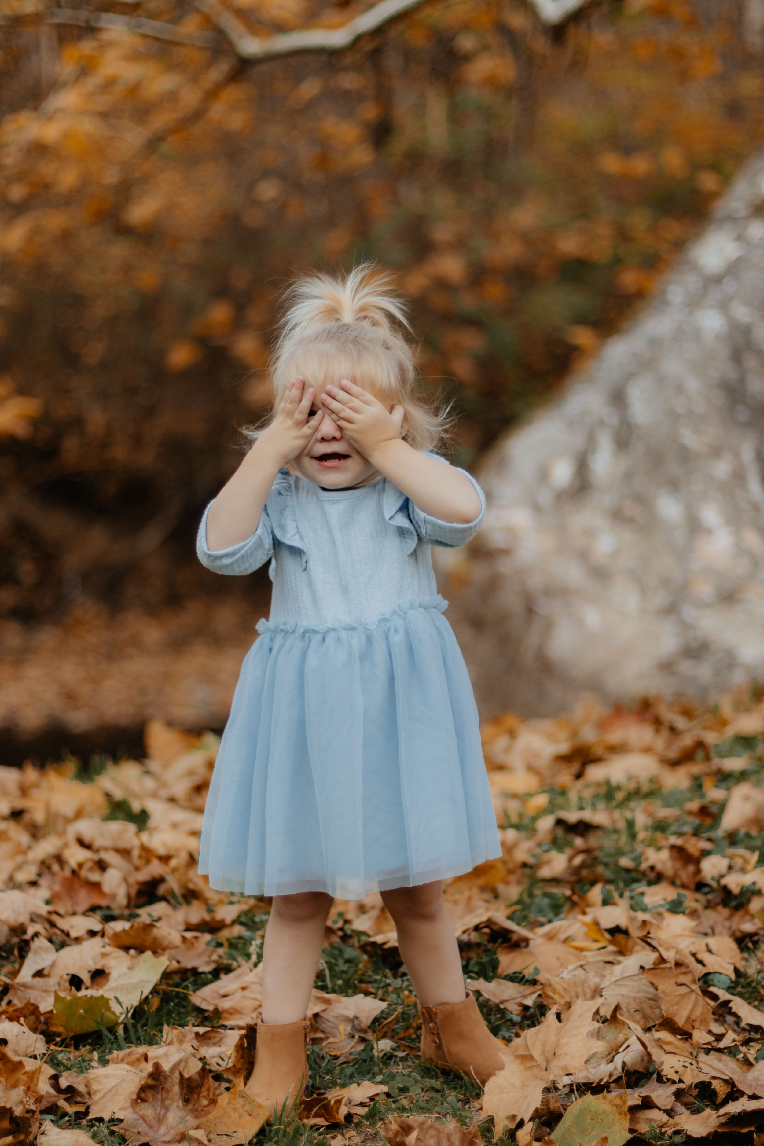 Peek-a-boo pose in blue dress surrounded by autumn leaves.