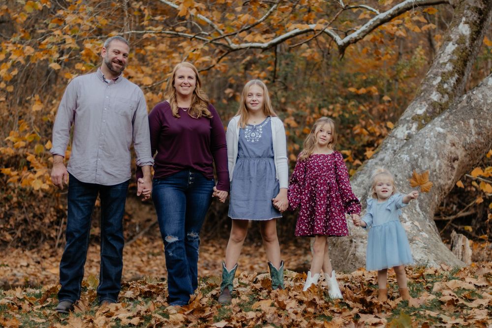 A family stands together in autumn foliage beneath a large tree branch with golden leaves in the background.