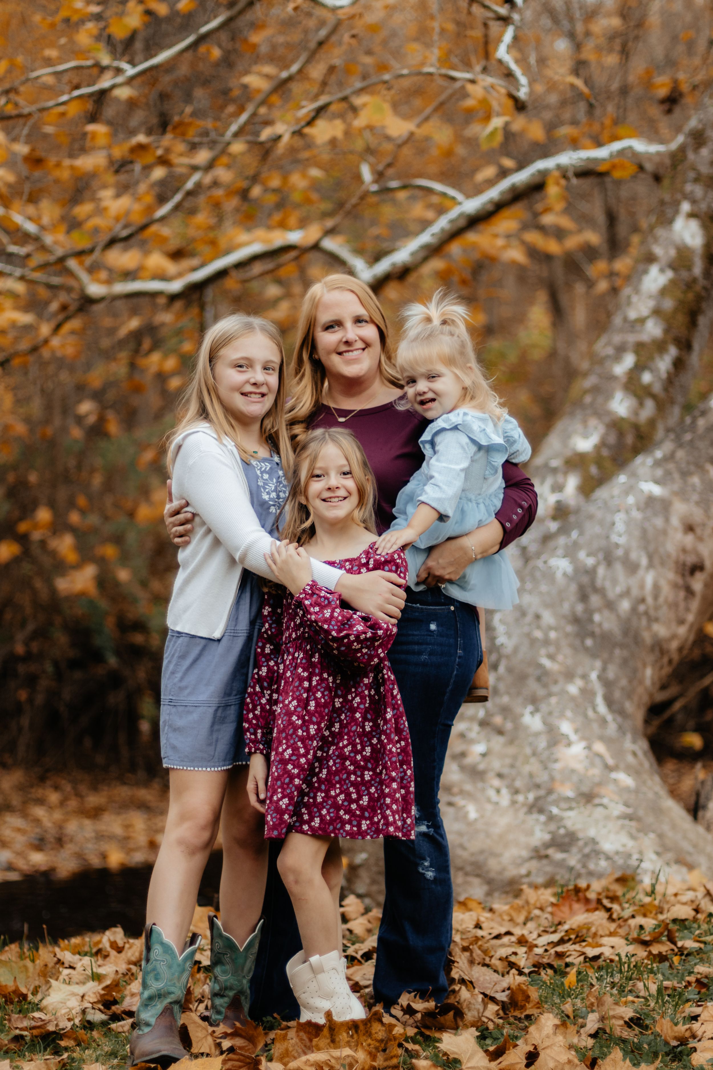 A group embraces while standing on fallen autumn leaves near a large tree.