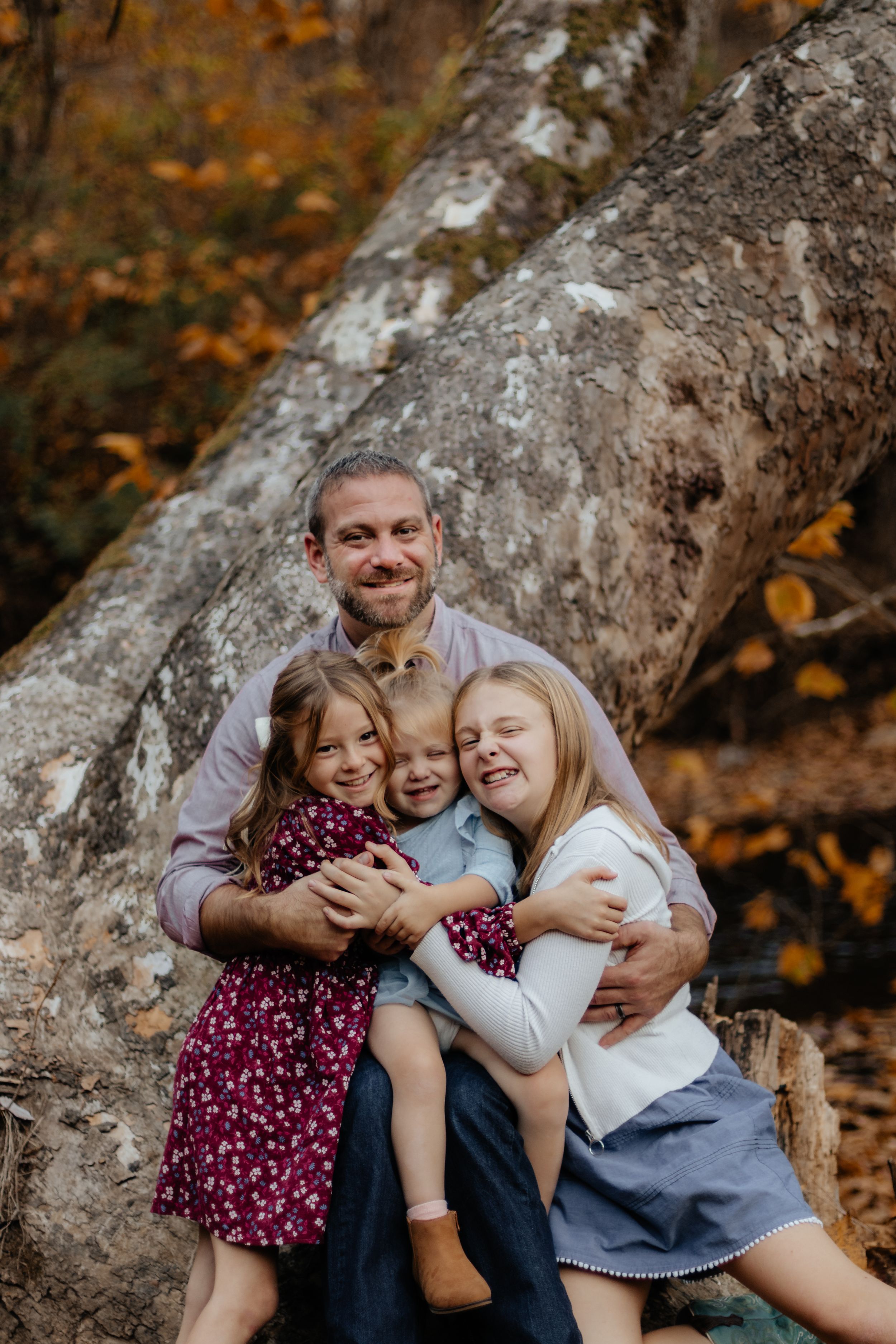 A heartwarming autumn family moment shared against a large tree trunk with golden fall foliage in the background.