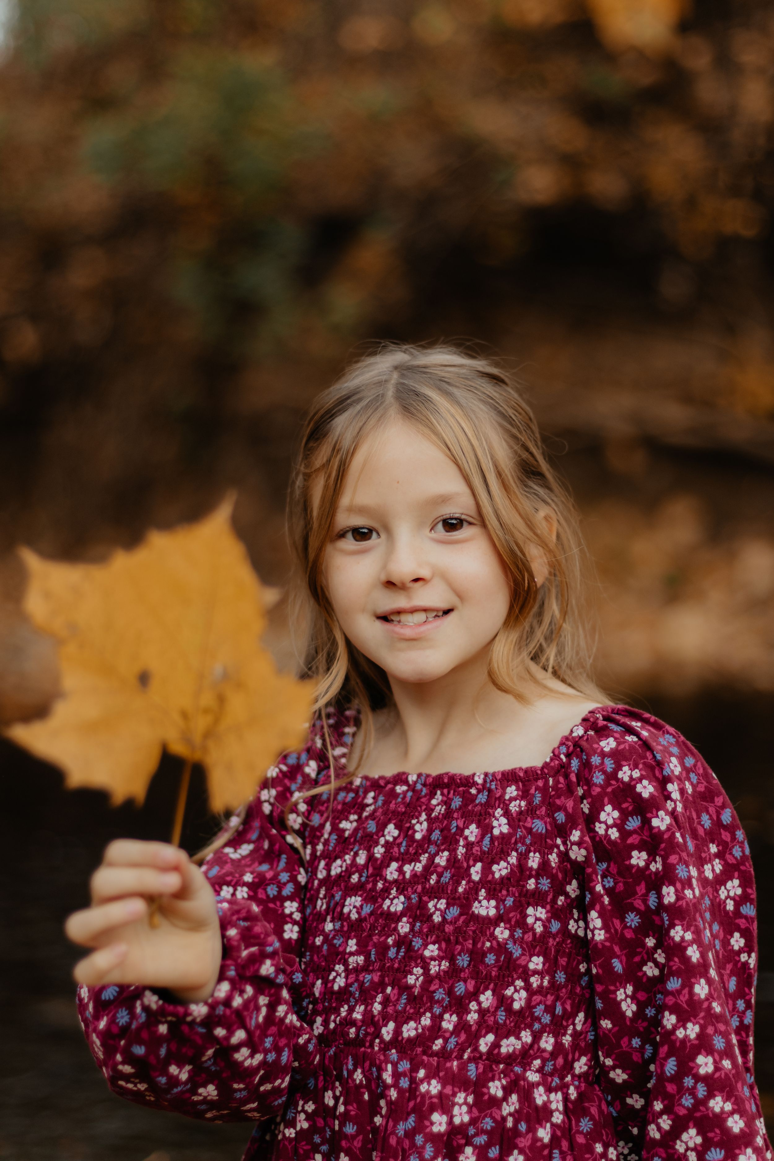 A child in a burgundy floral dress holds up yellow autumn leaves while playing outdoors.