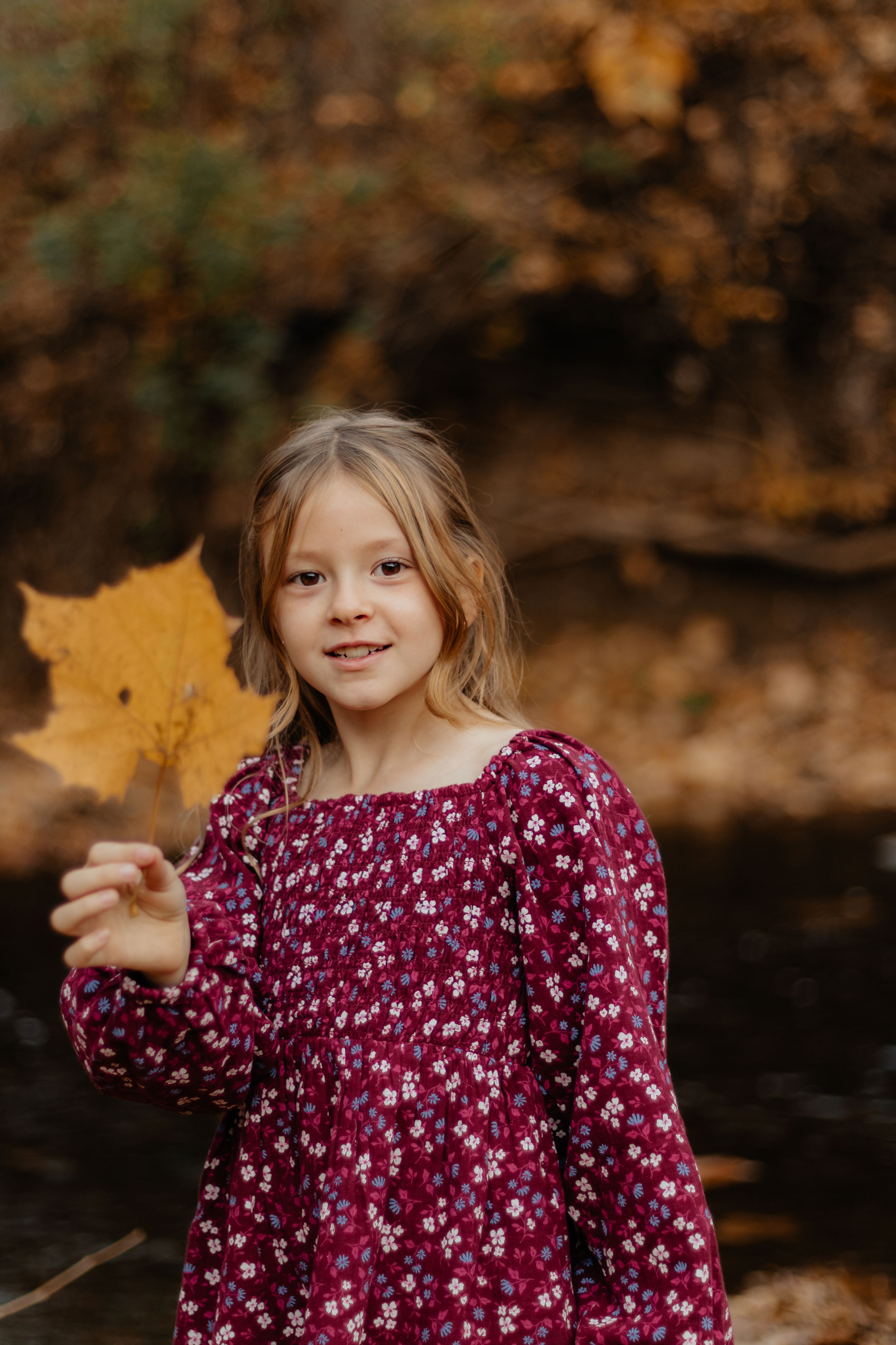 A child wearing a burgundy floral dress holds up a large yellow maple leaf in an autumn setting.