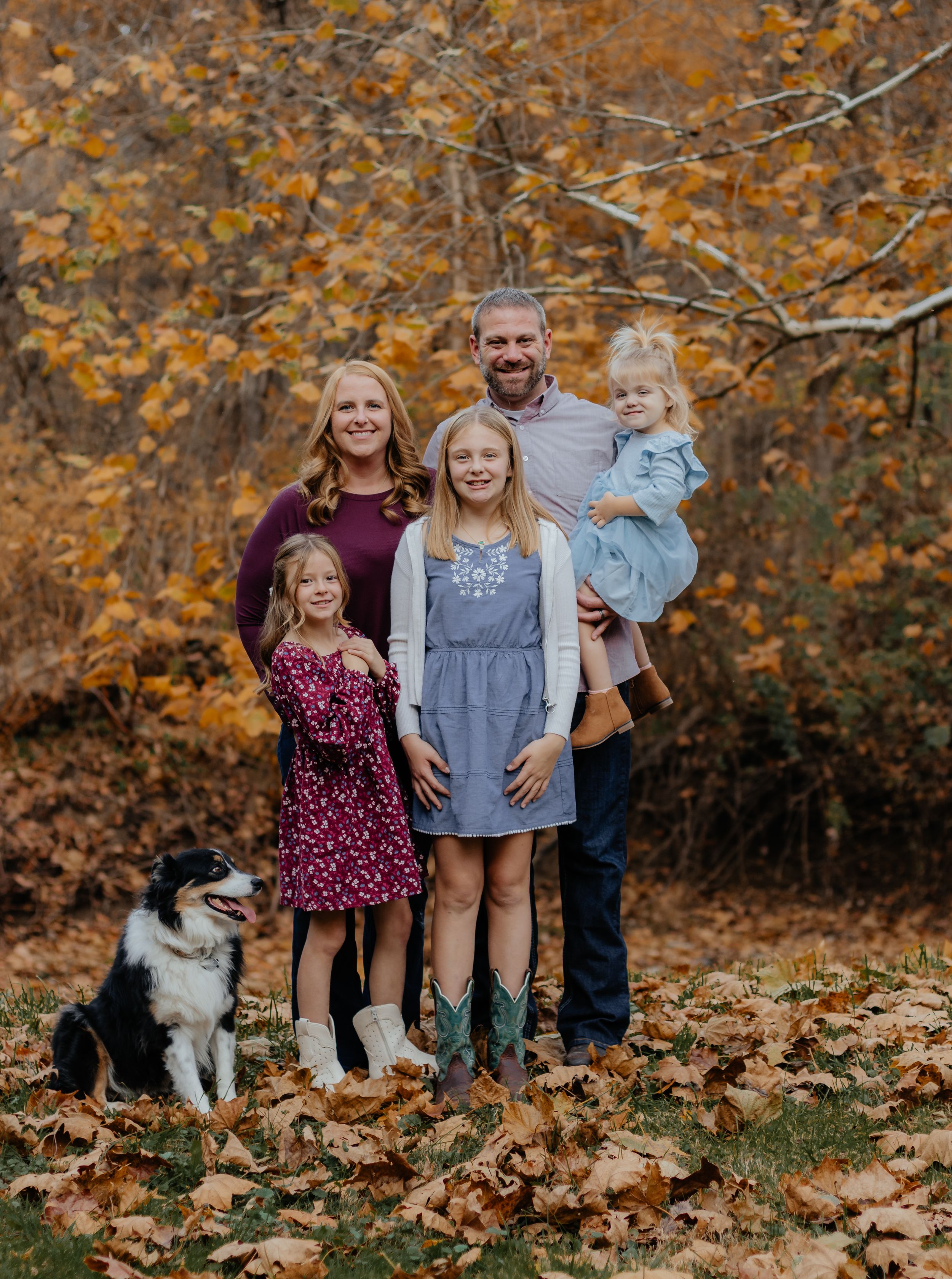 Family and dog posing for photo on autumn leaves with orange foliage background.