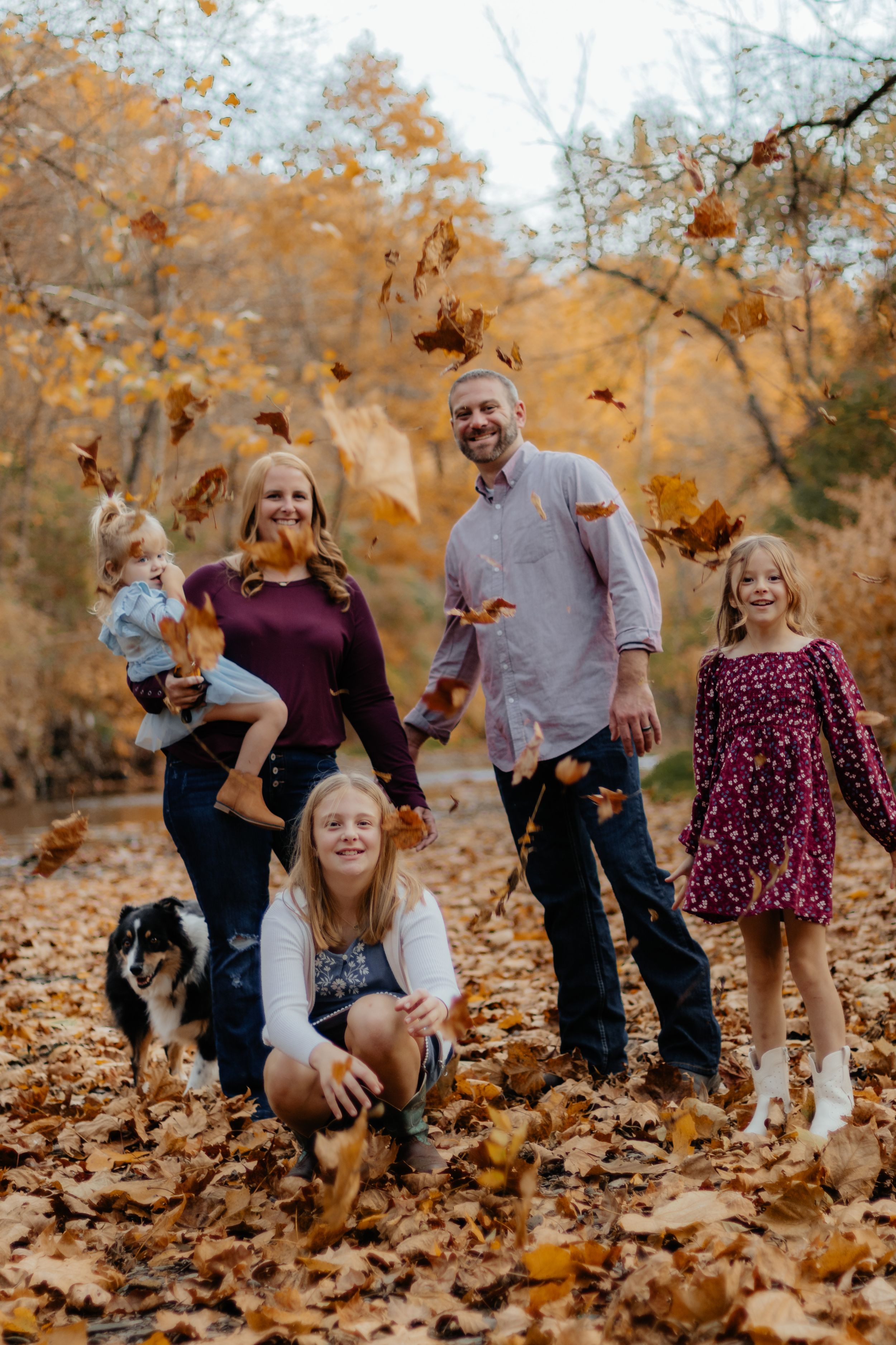 Family members laugh and play in piles of fallen autumn leaves during an outdoor photography session.